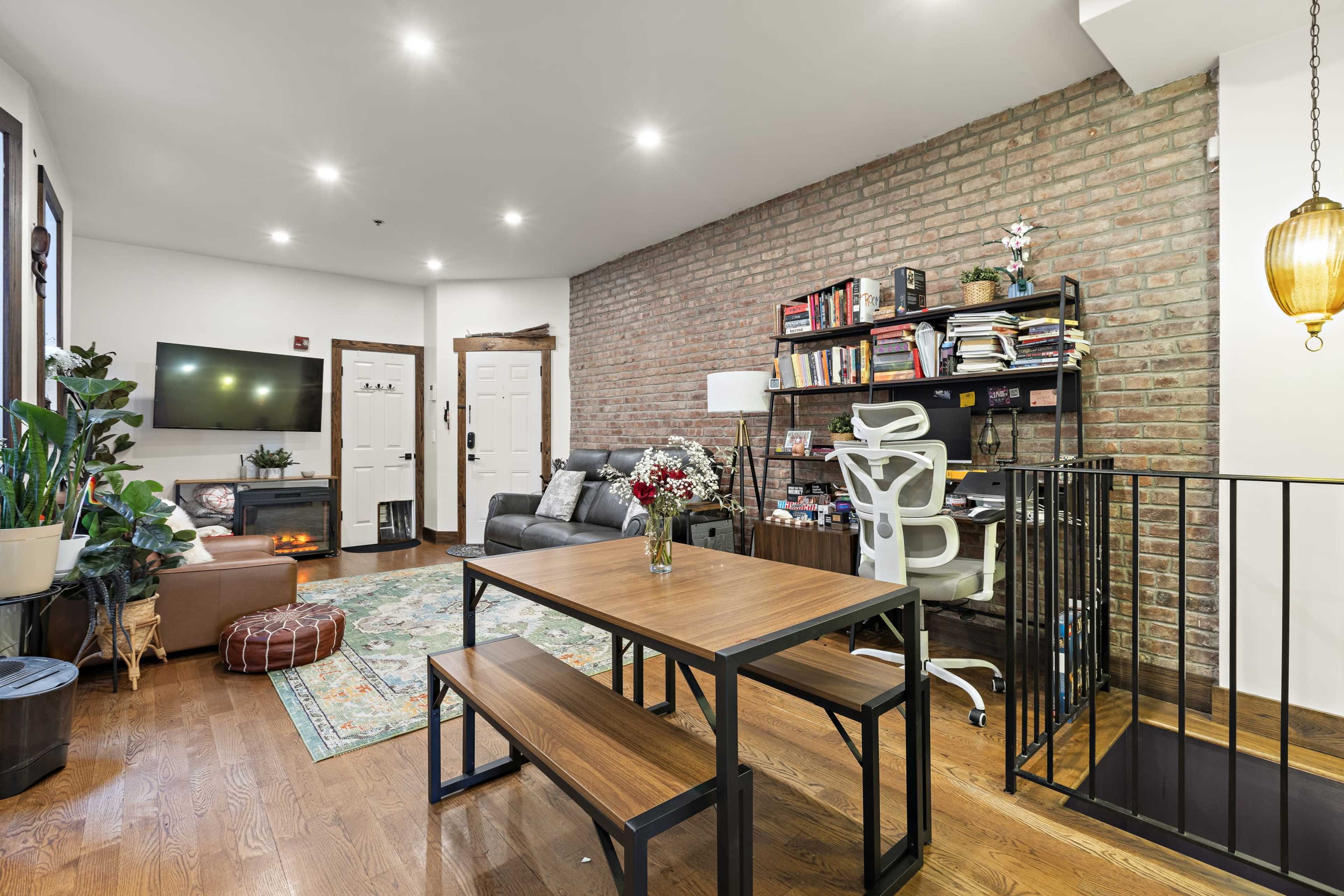 The image shows a cozy living space featuring a seating area with a sofa, a dining table, a work desk, and a mix of greenery and decorative items against a brick wall.