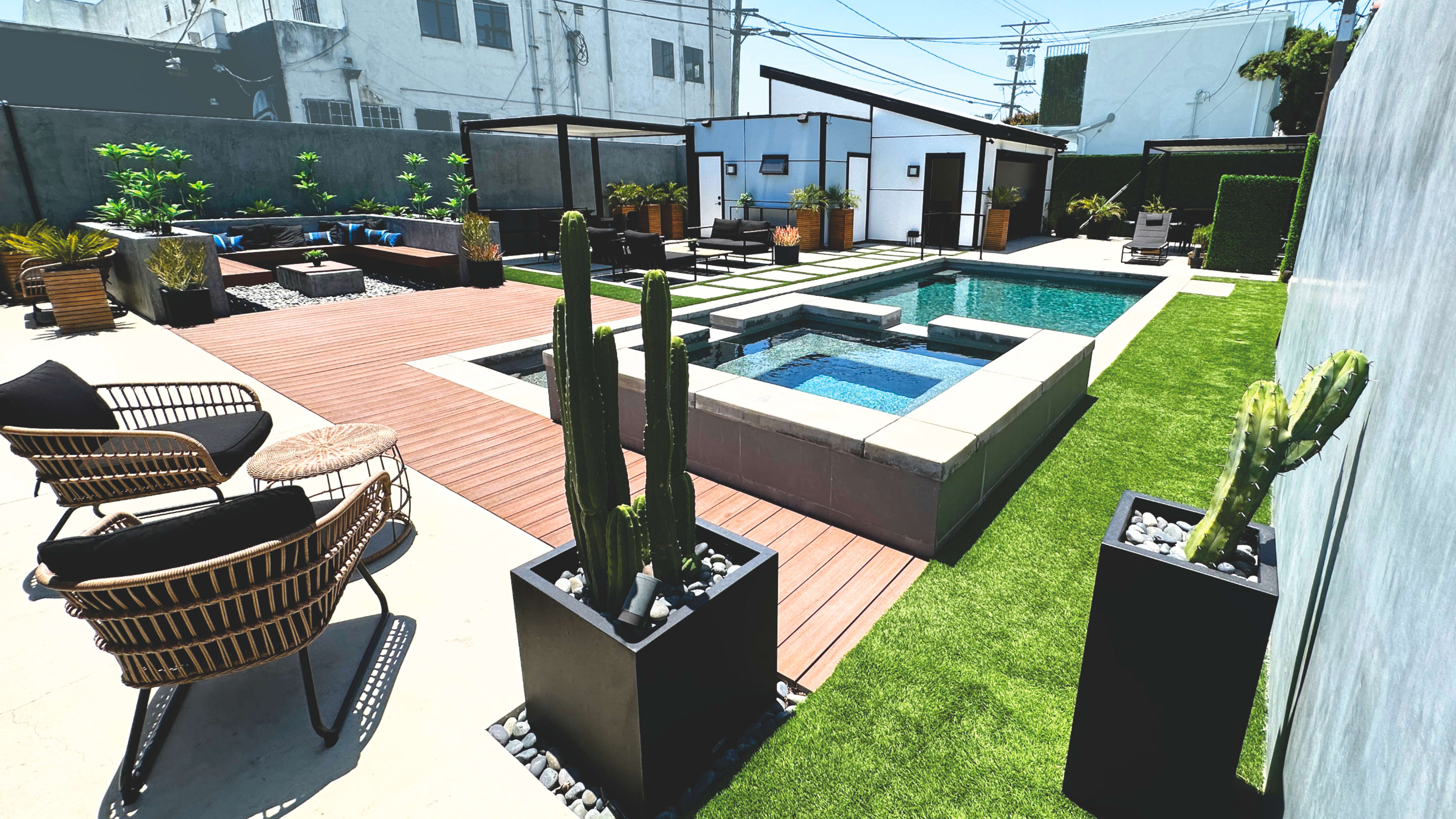 The image shows a contemporary backyard featuring a swimming pool, spa area, wooden decking, landscaped plants, and outdoor seating arrangements.