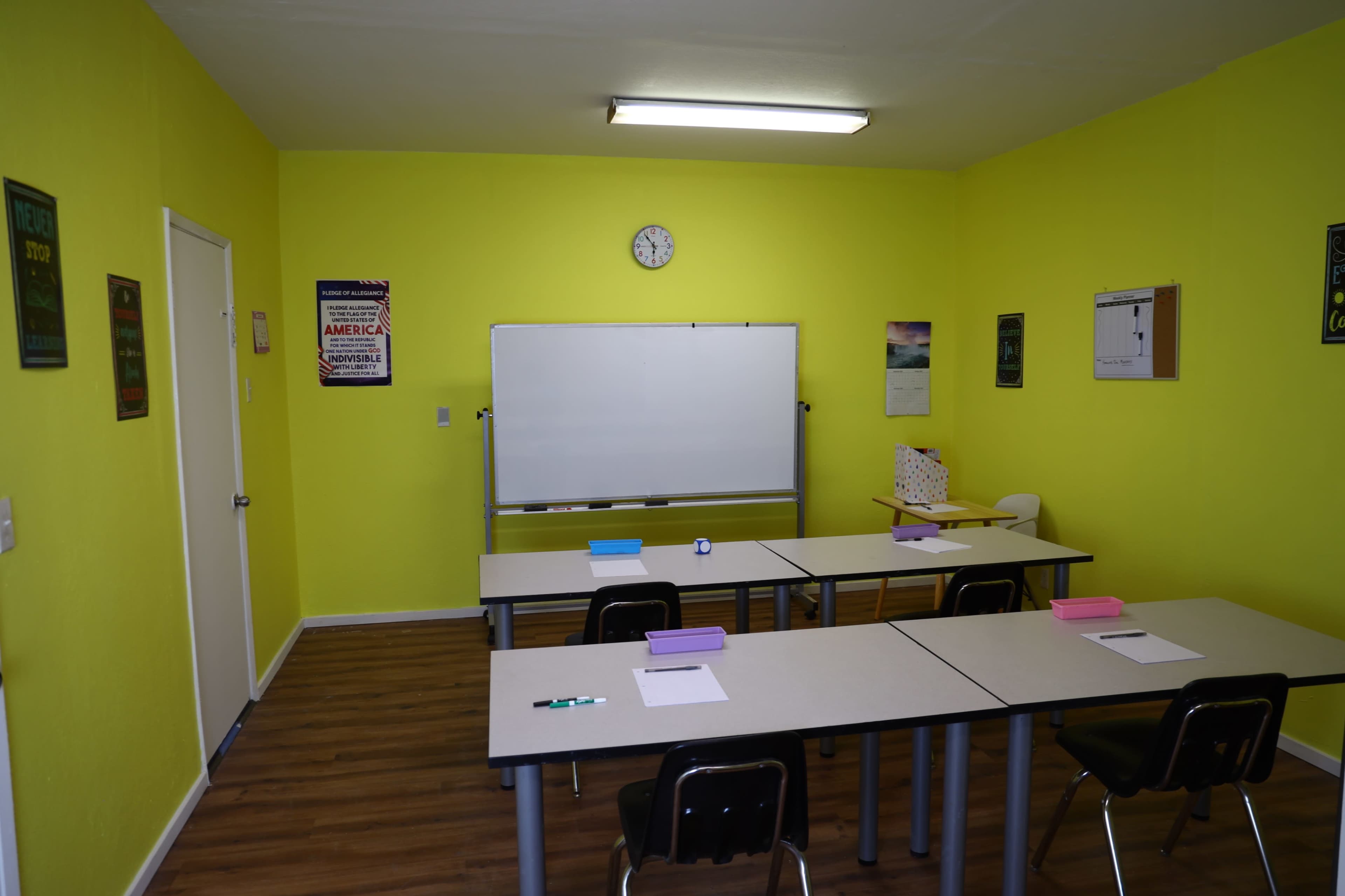A brightly colored classroom features several desks arranged in rows, a whiteboard at the front, and a clock on the wall.