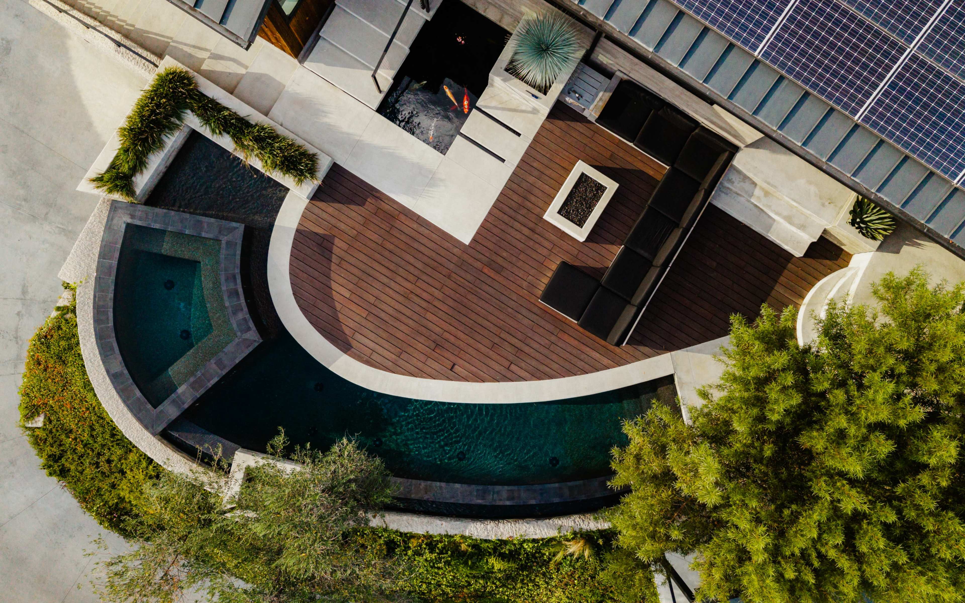 The image shows an aerial view of a modern outdoor patio with a wooden deck, a water feature, and surrounding greenery.