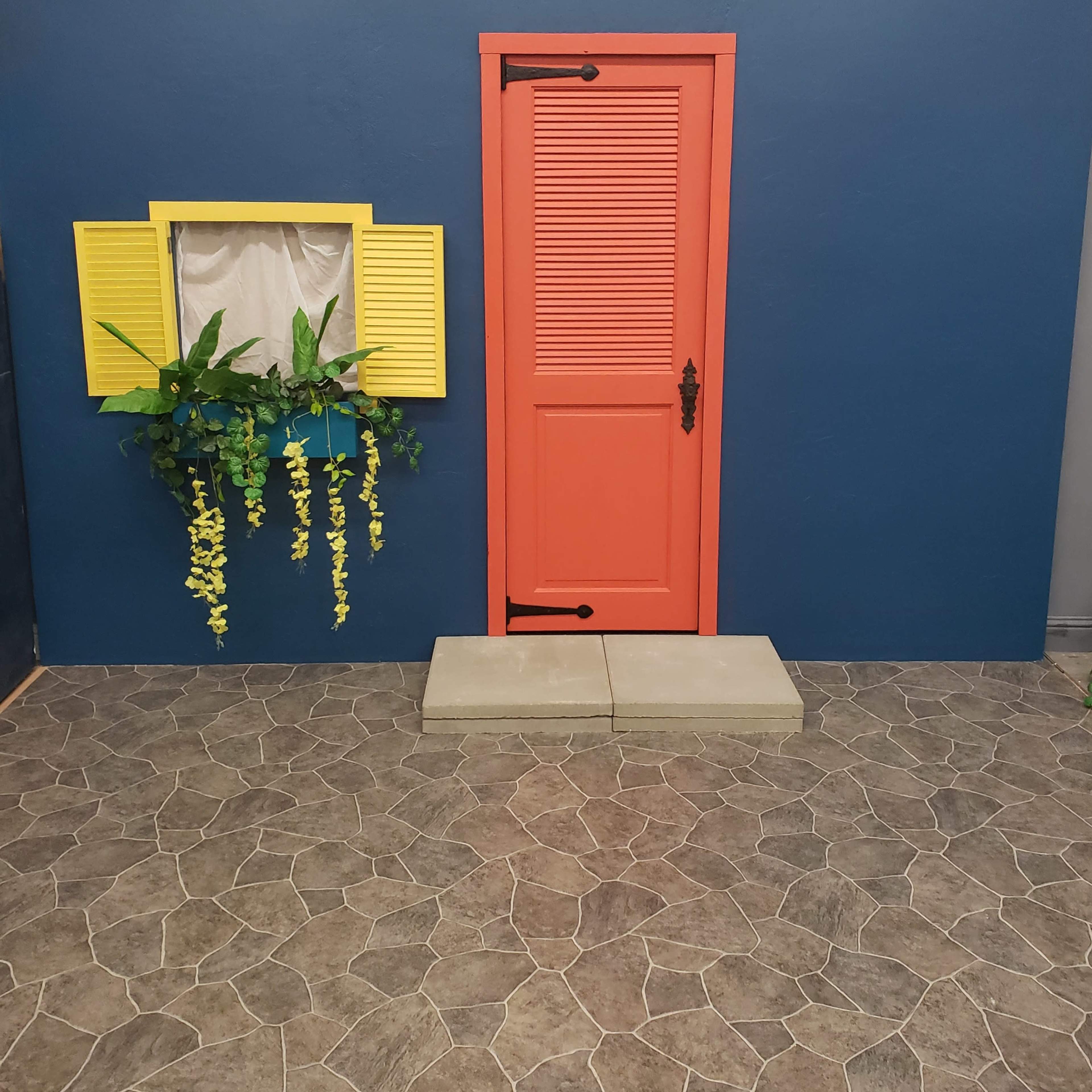 The scene features a brightly colored coral door and a yellow window with shutters, both set against a blue wall, complemented by a green planter and a stone-patterned floor.