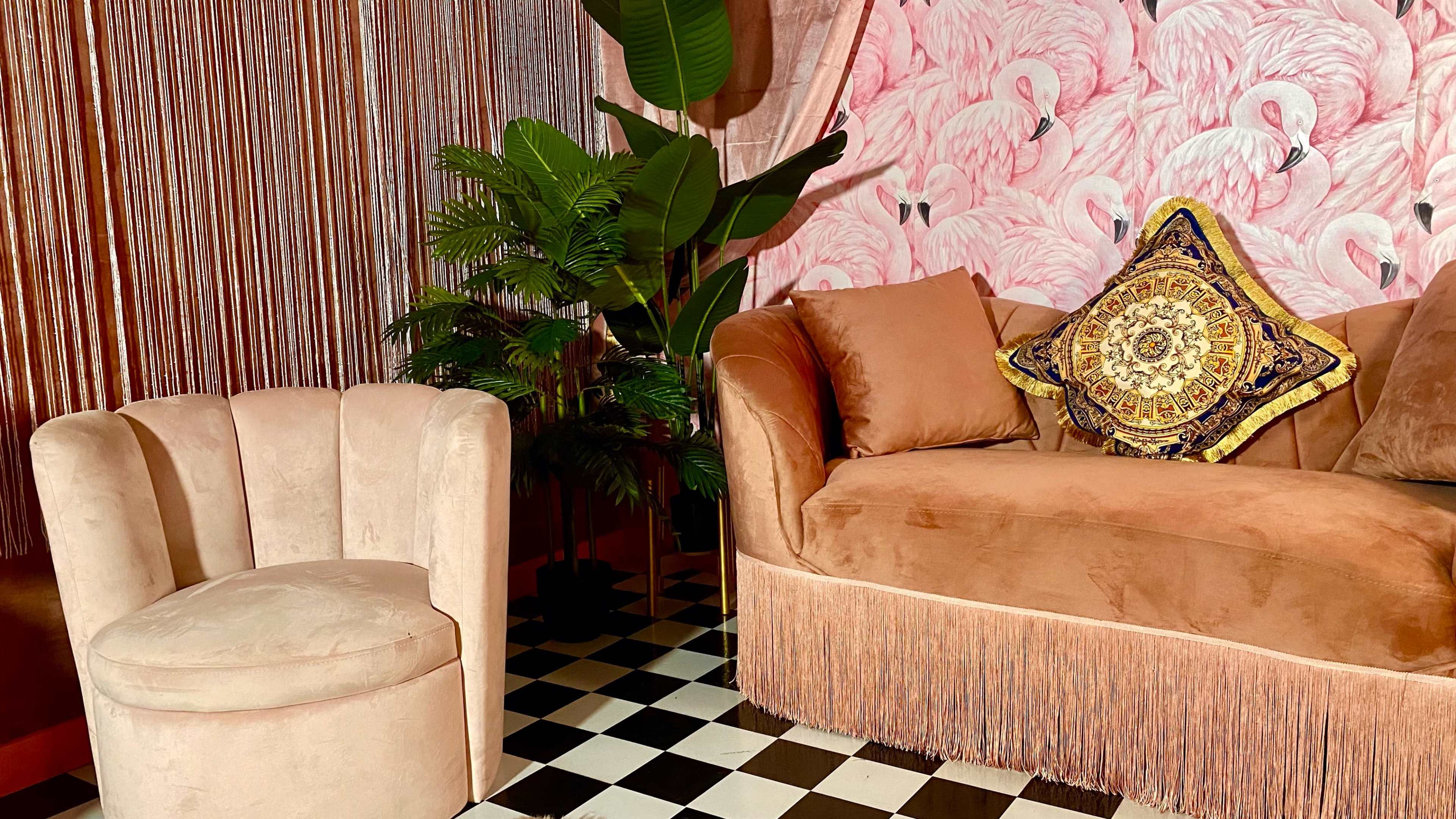 A plush couch with fringe and an ornate pillow is paired with a small upholstered chair, set against a pink flamingo wallpaper and surrounded by indoor plants.