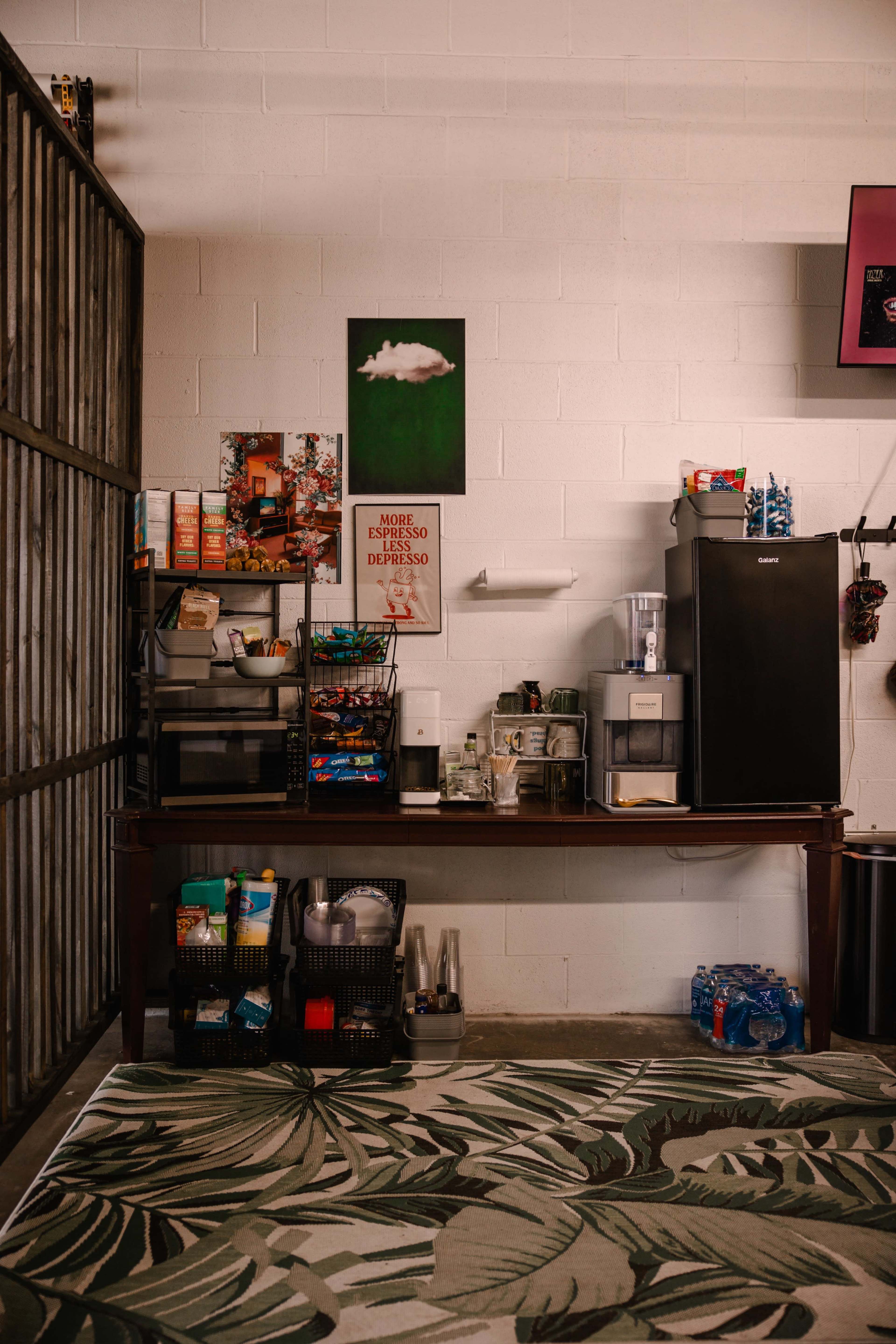 A workspace with a large table featuring various kitchen supplies, a coffee maker, and a refrigerator against a wall decorated with a green artwork and a motivational poster.