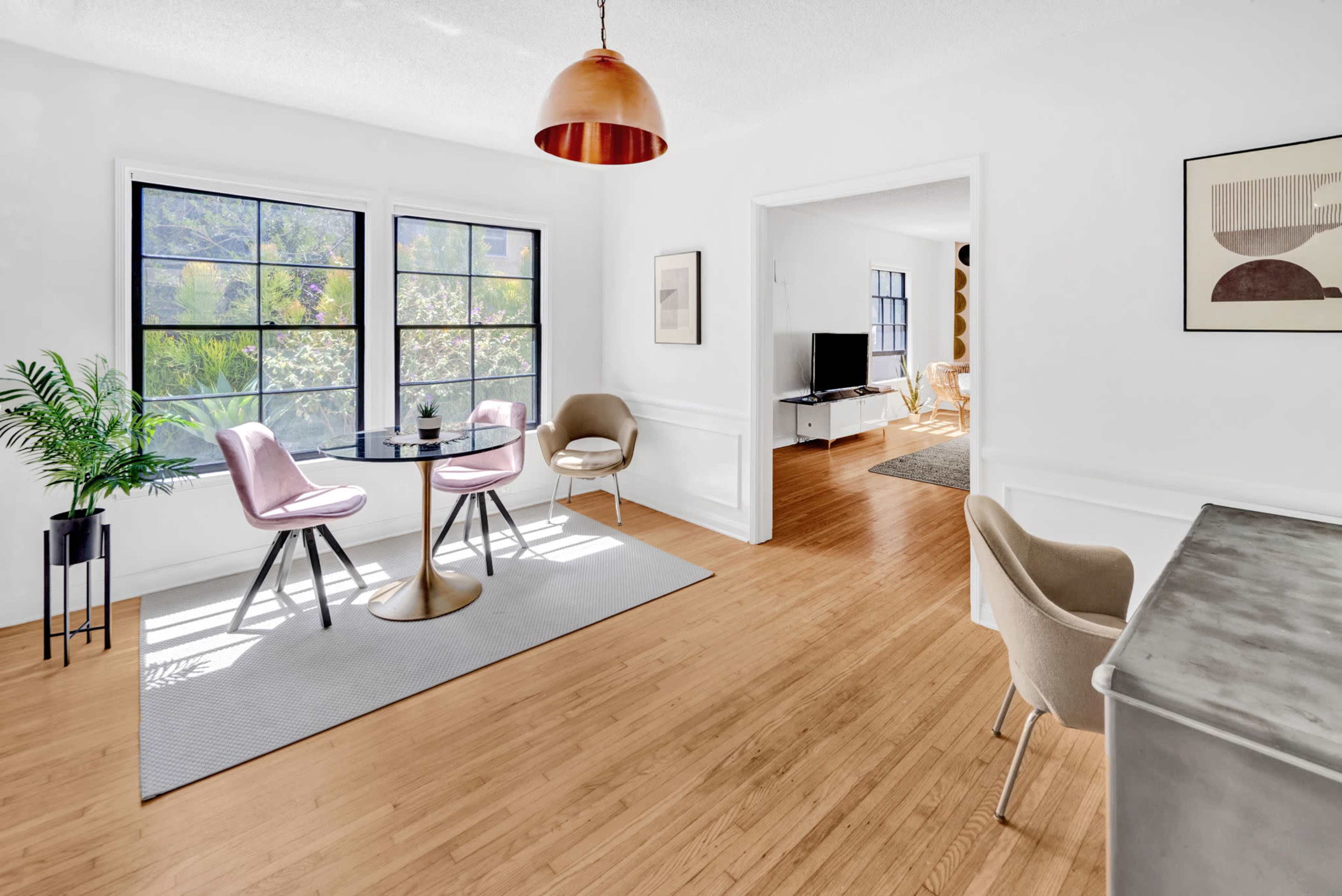 A bright, modern living space features a round dining table with four chairs, large windows, and a view of greenery.