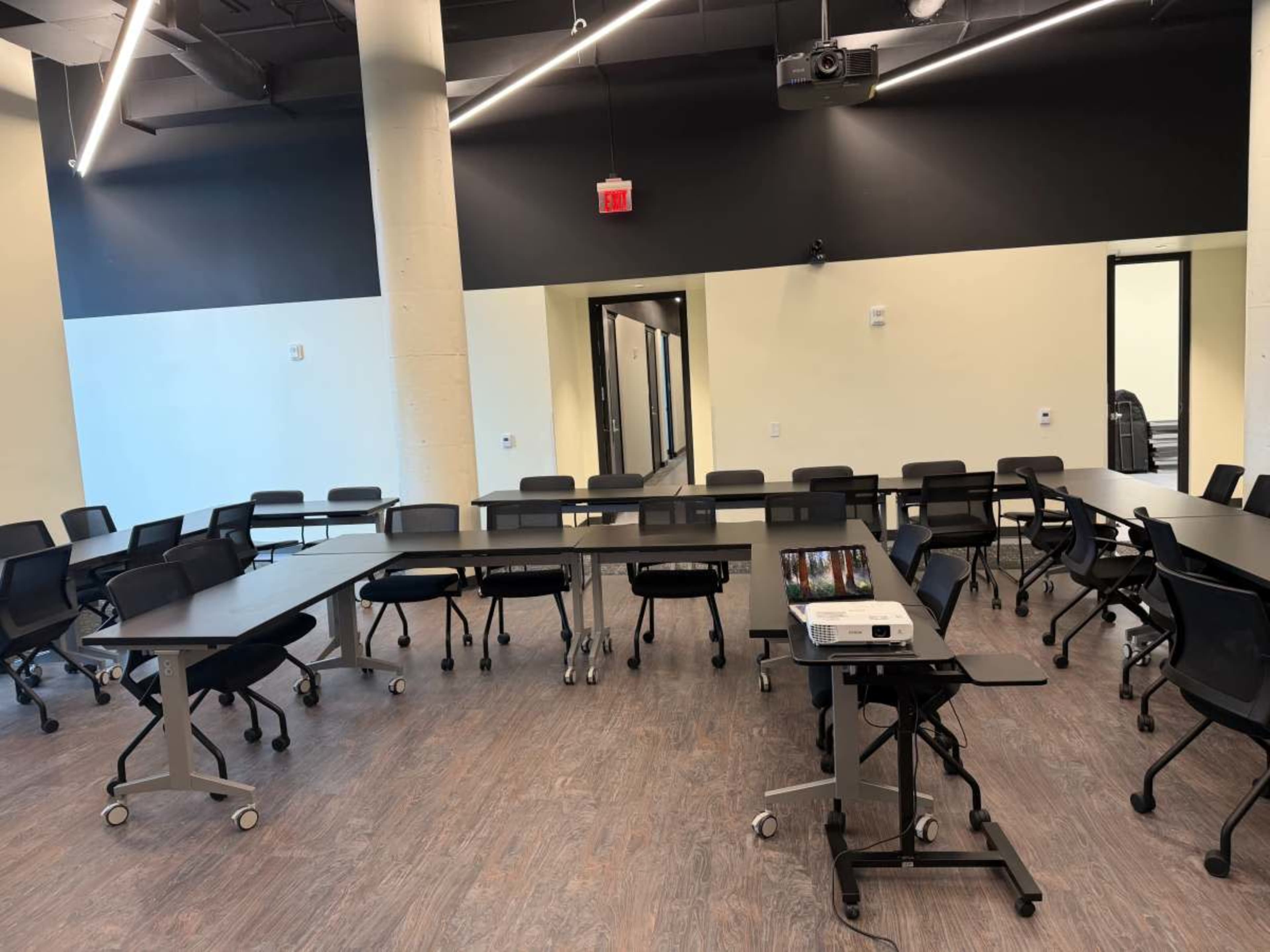The image shows a conference room set up with several tables arranged in a U-shape, surrounded by black chairs and equipped with a projector.