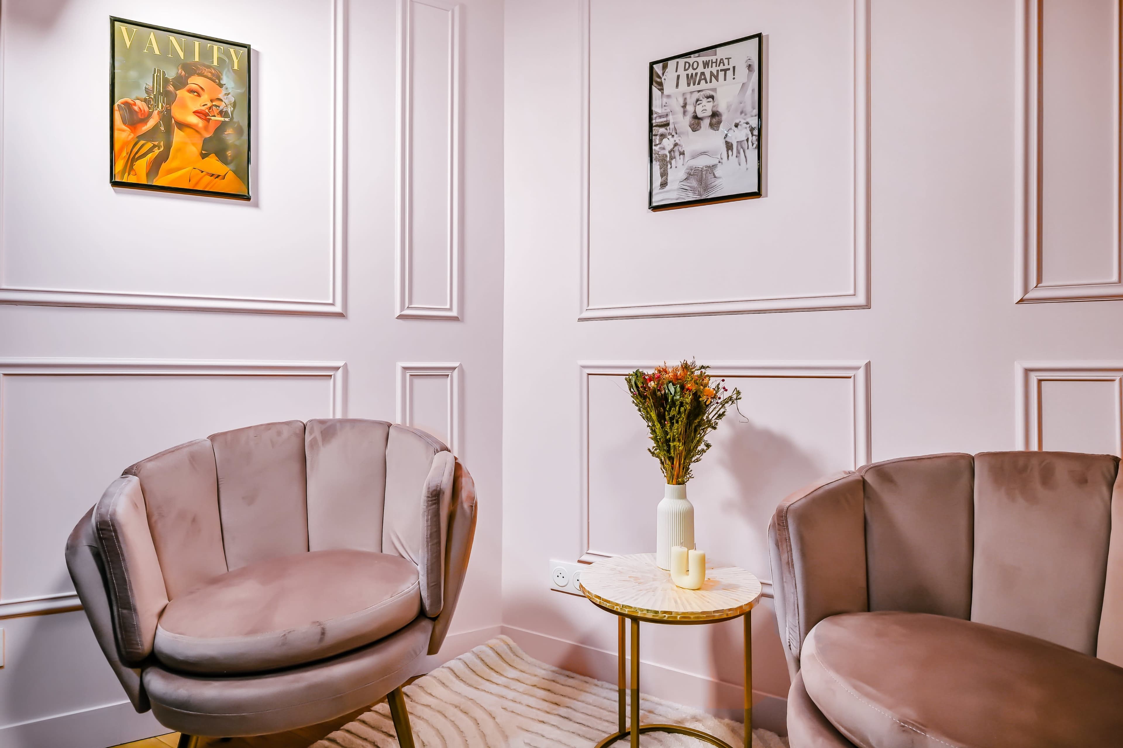 The image shows a small corner of a room featuring two plush, round chairs beside a gold side table topped with a vase of flowers, against a pale pink wall adorned with framed artwork.