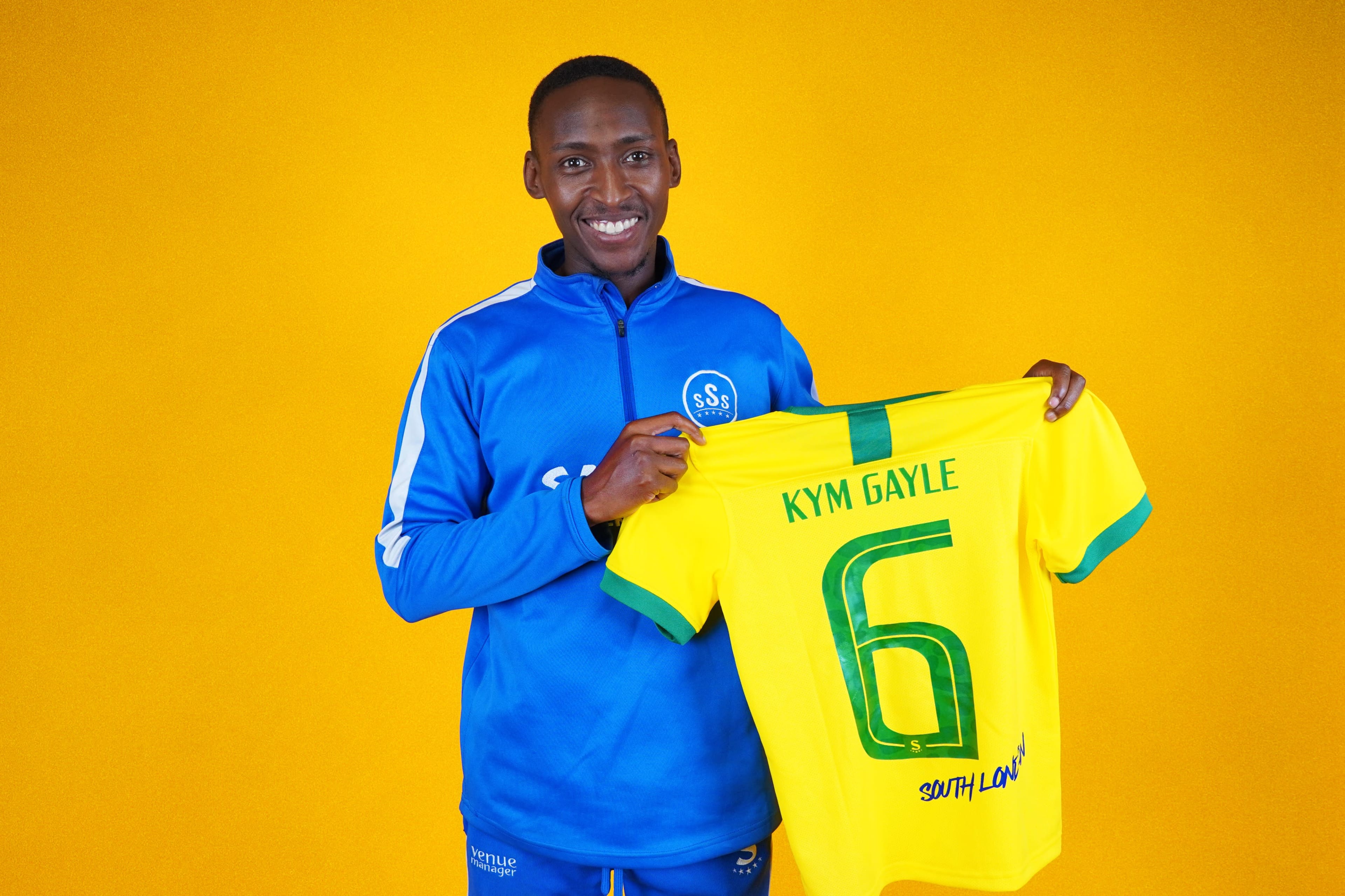 A person in a blue jacket is smiling while holding up a yellow sports jersey with the name "KYM GAYLE" and the number "6" printed on it, against a bright yellow background.