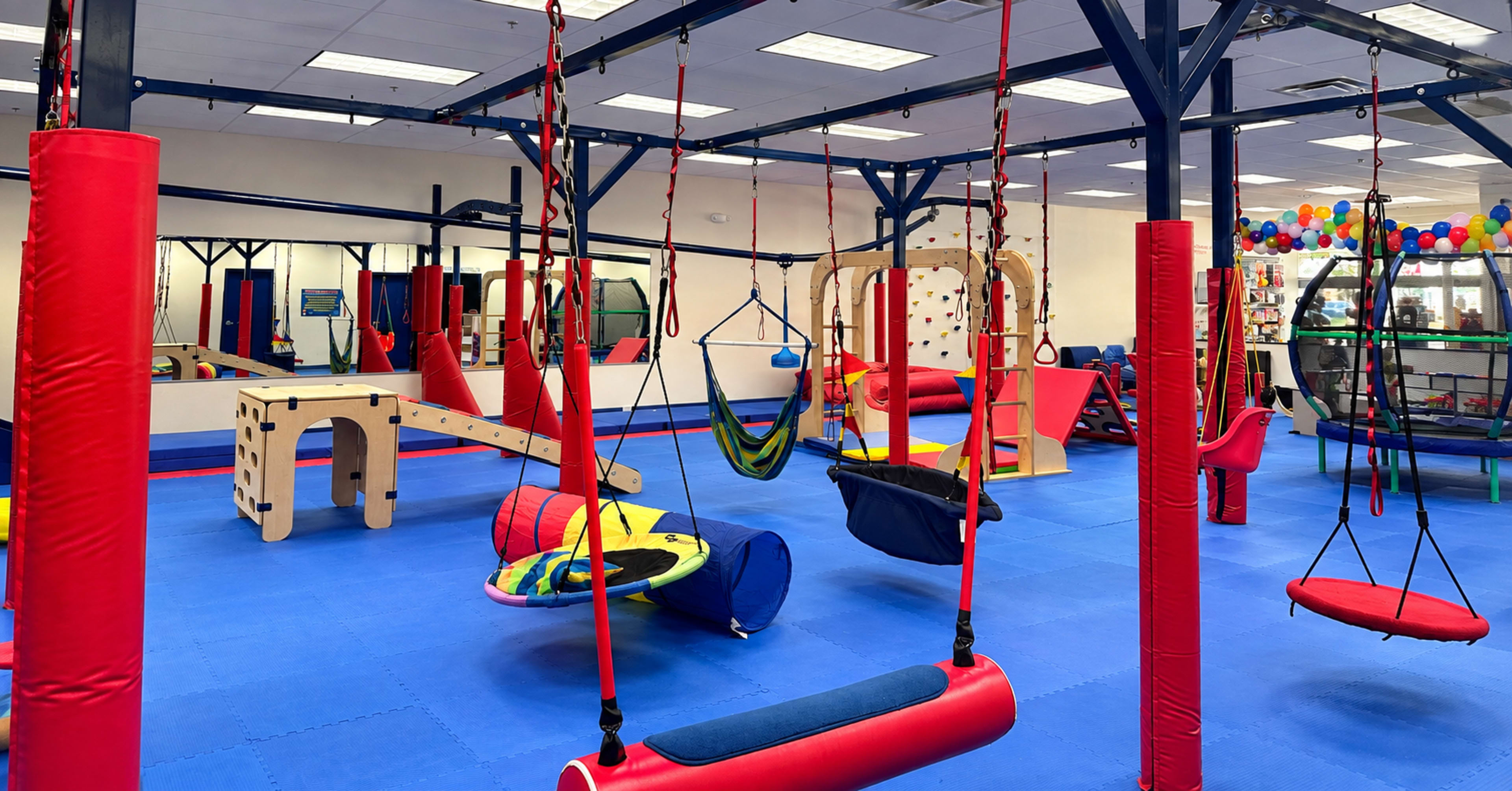 The image shows an indoor play area featuring various colorful gym equipment, including swings, a hammock, and obstacles, all set on a blue rubberized floor.