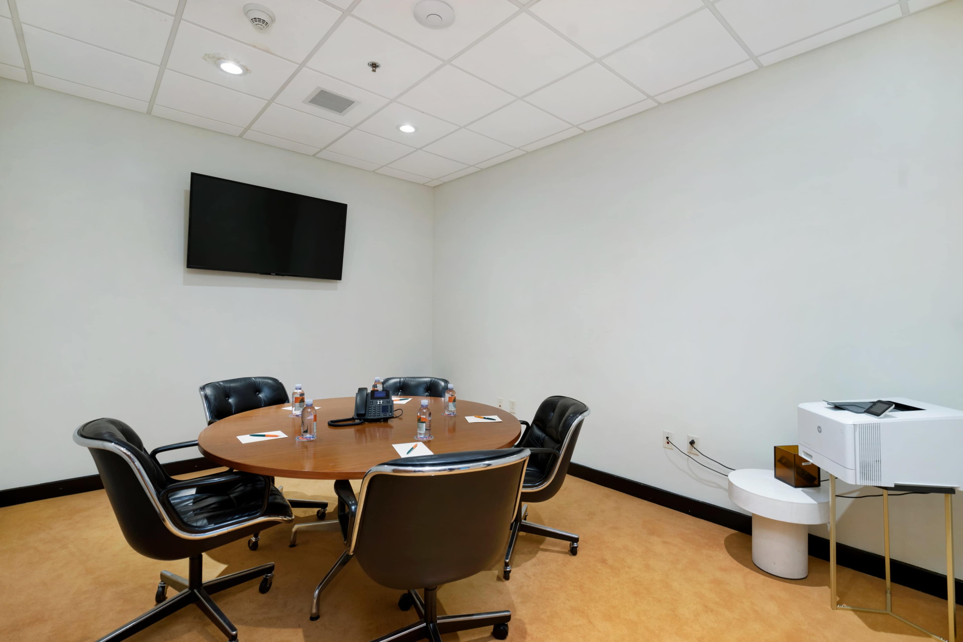 A small conference room features a round wooden table surrounded by four black leather chairs, a wall-mounted TV, a printer, and water bottles on the table.