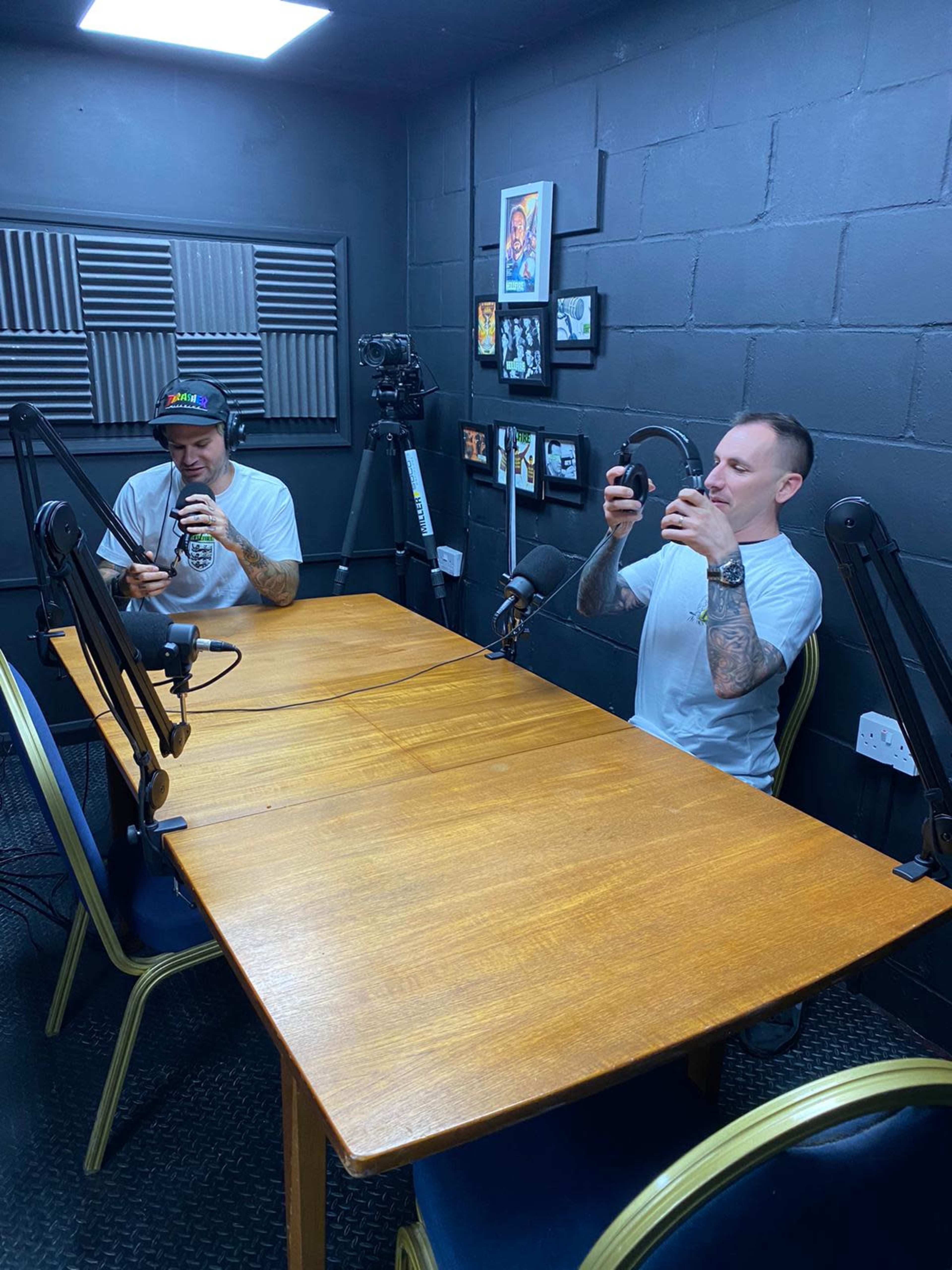 Two individuals sit at a wooden table in a recording studio equipped with microphones and soundproofing panels.