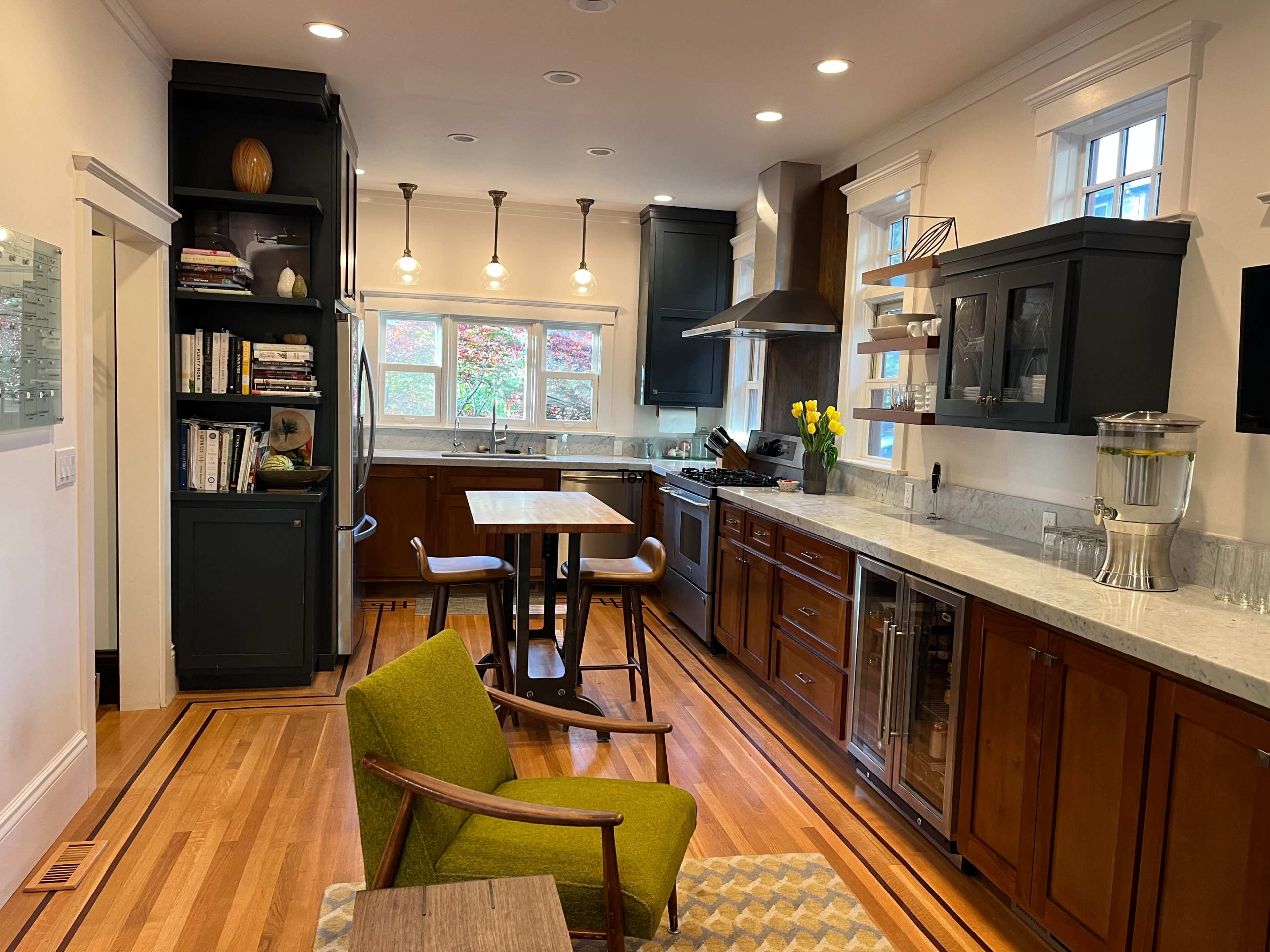 A modern kitchen features dark wood cabinets, a marble countertop, and a small dining table, with ample natural light from windows.