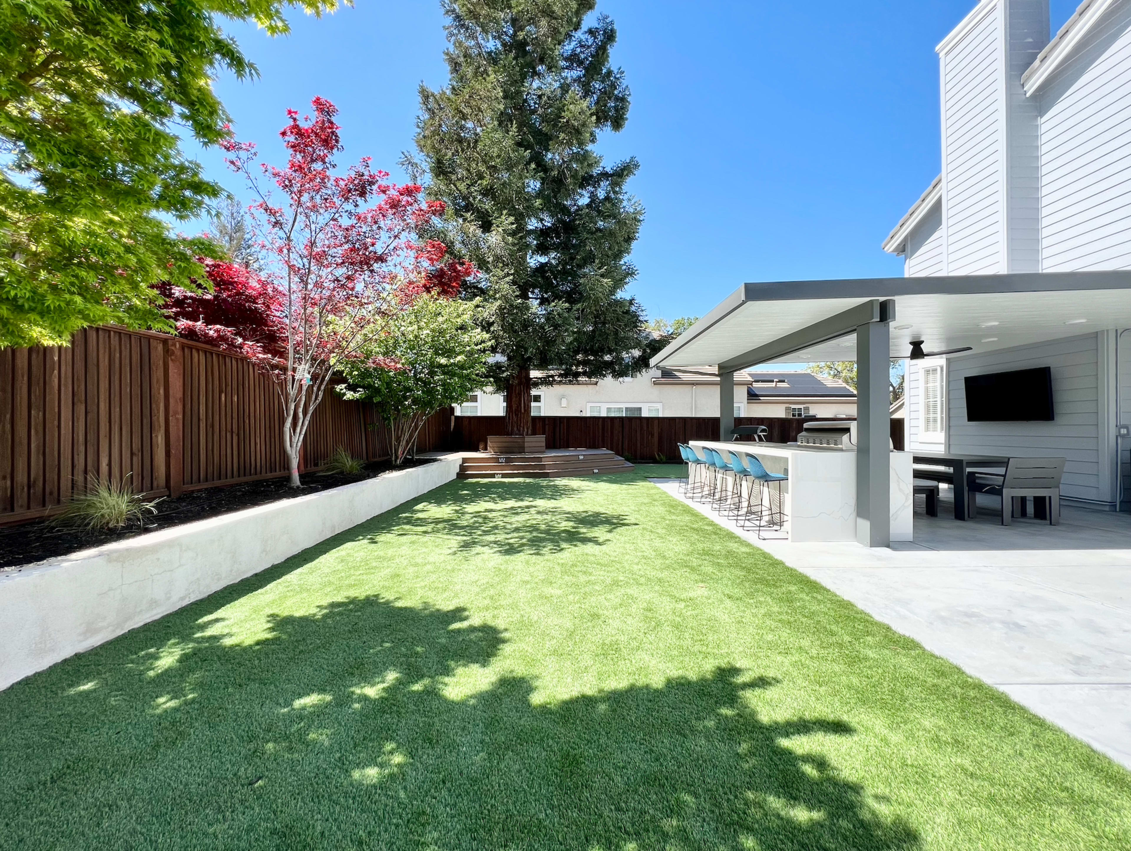 A landscaped backyard featuring artificial grass, a dining area with bar stools, and several trees along a wooden fence.