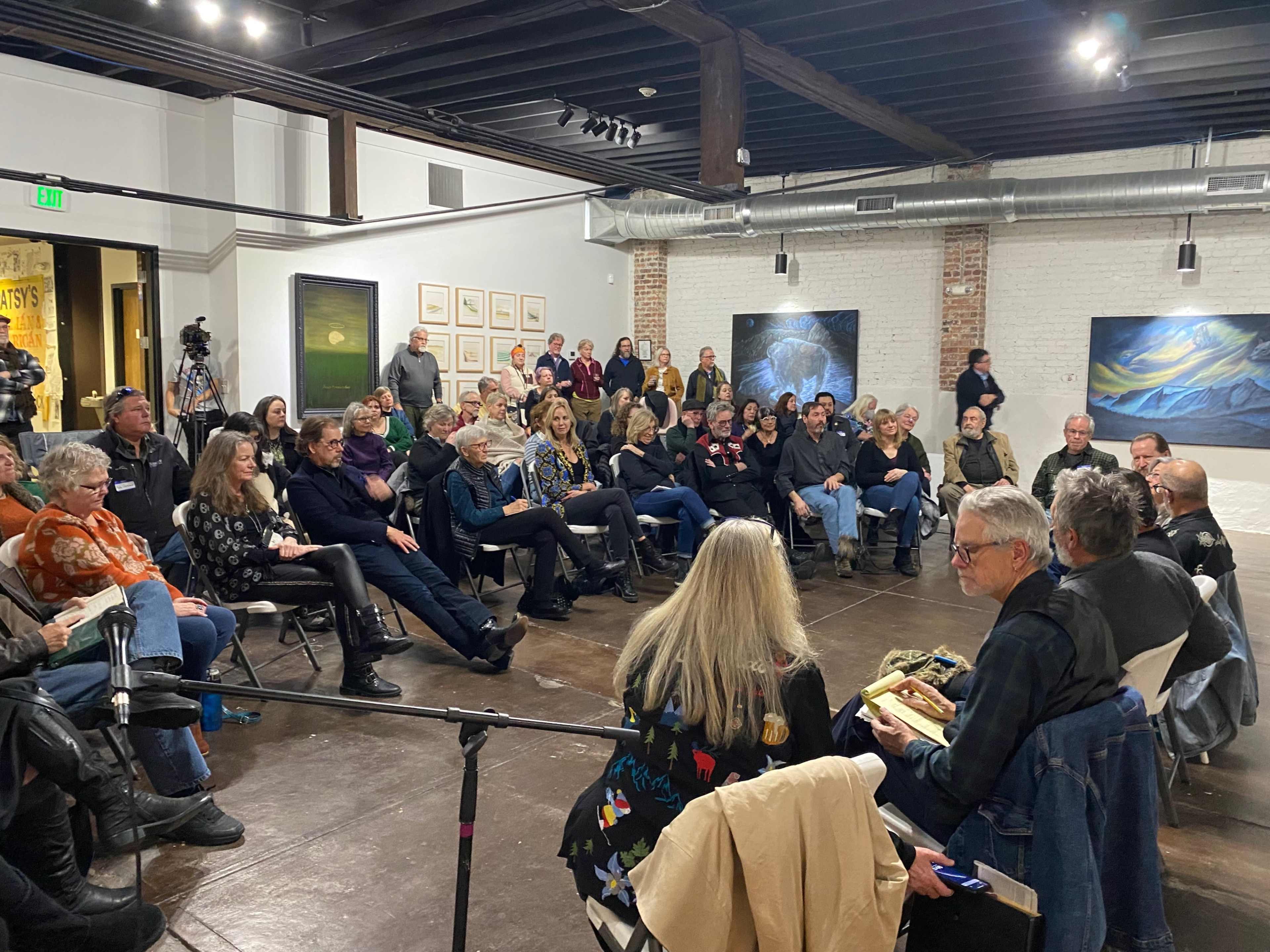 A crowd of people is seated in a gallery space, attentively listening to speakers at a discussion event.