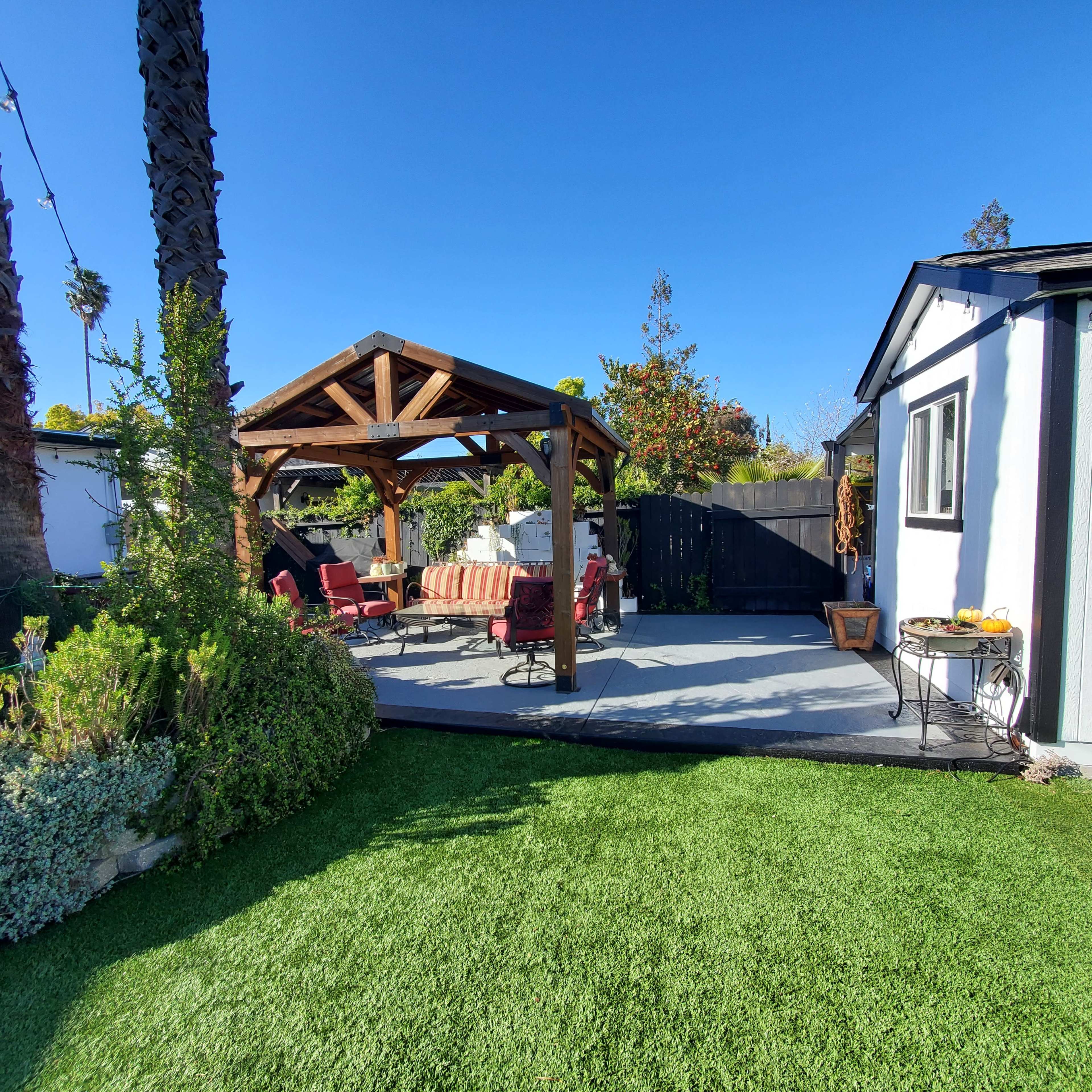 A wooden pergola with red chairs is set on a patio surrounded by greenery and a black fence.
