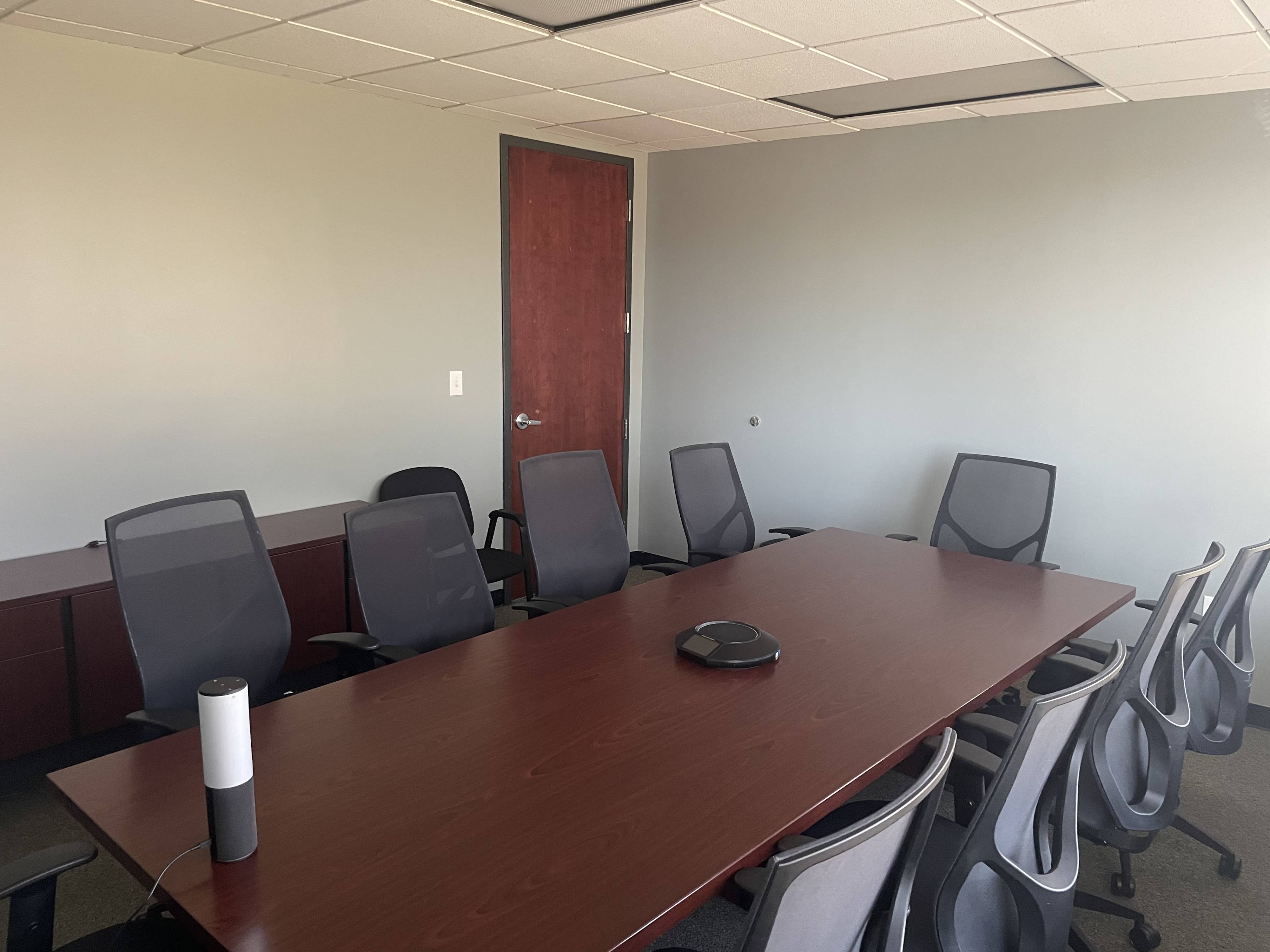 A conference room with a large wooden table surrounded by several office chairs and a speakerphone in the center.