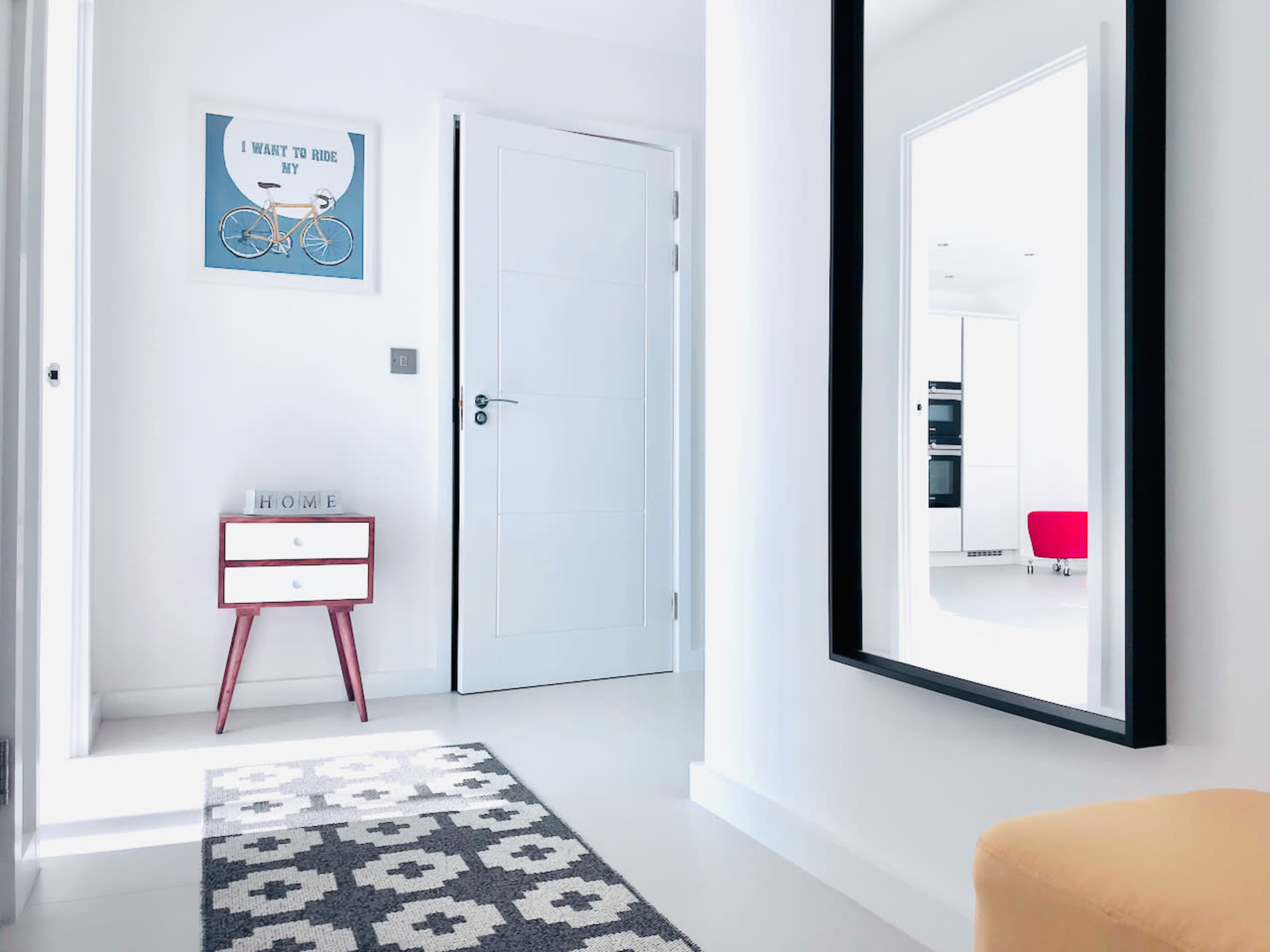 A bright hallway features a bicycle-themed poster, a small white drawer with pink legs, and a large mirror reflecting an adjacent room.