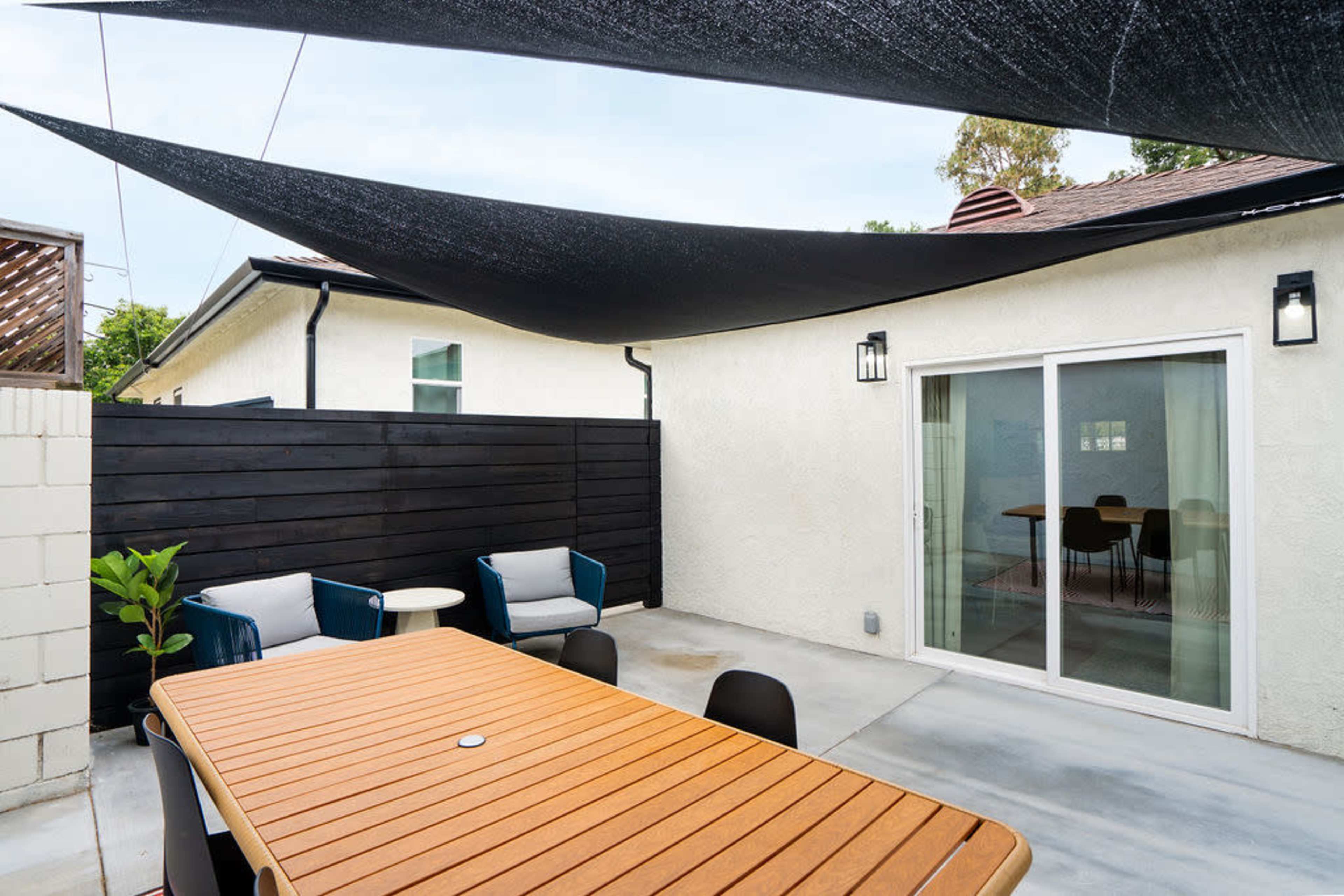 A modern outdoor space features a wooden table surrounded by chairs, shaded by black canopies, with sliding glass doors leading into a light-colored interior room.