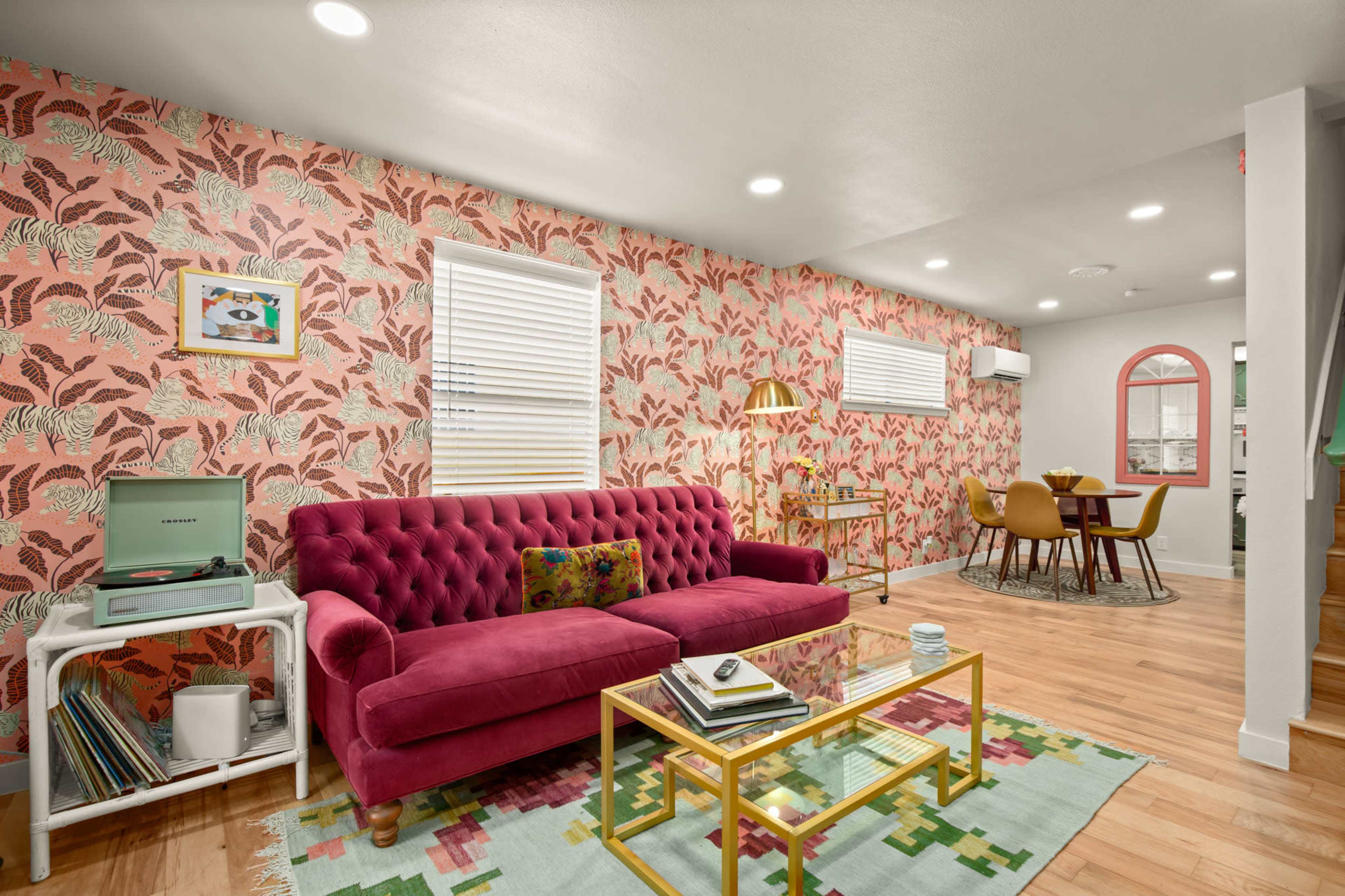 The image shows a stylish living room with a pink tufted sofa, patterned wallpaper, a coffee table, and a dining area with a round table and chairs.