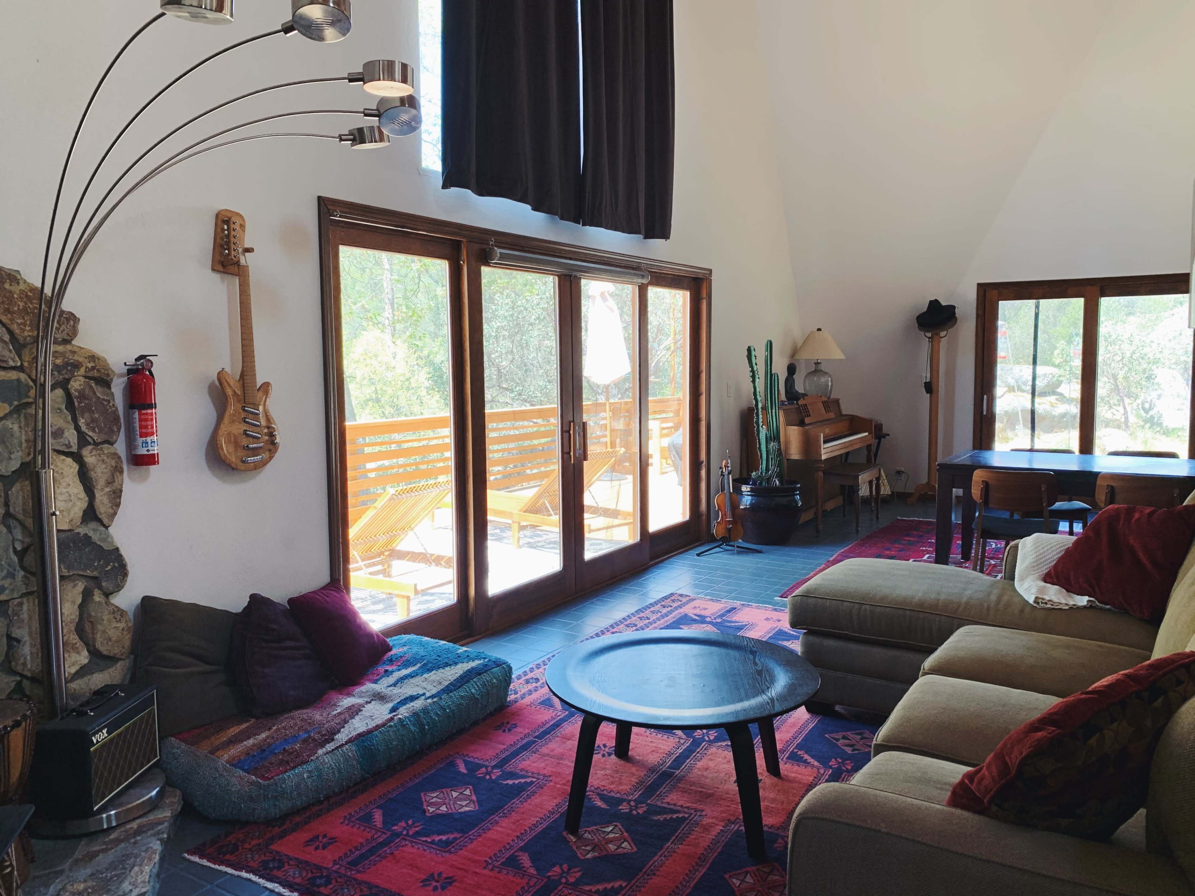 The interior of a spacious living room features large windows overlooking a deck, a guitar mounted on the wall, and a mix of seating options including a sofa and floor cushions.