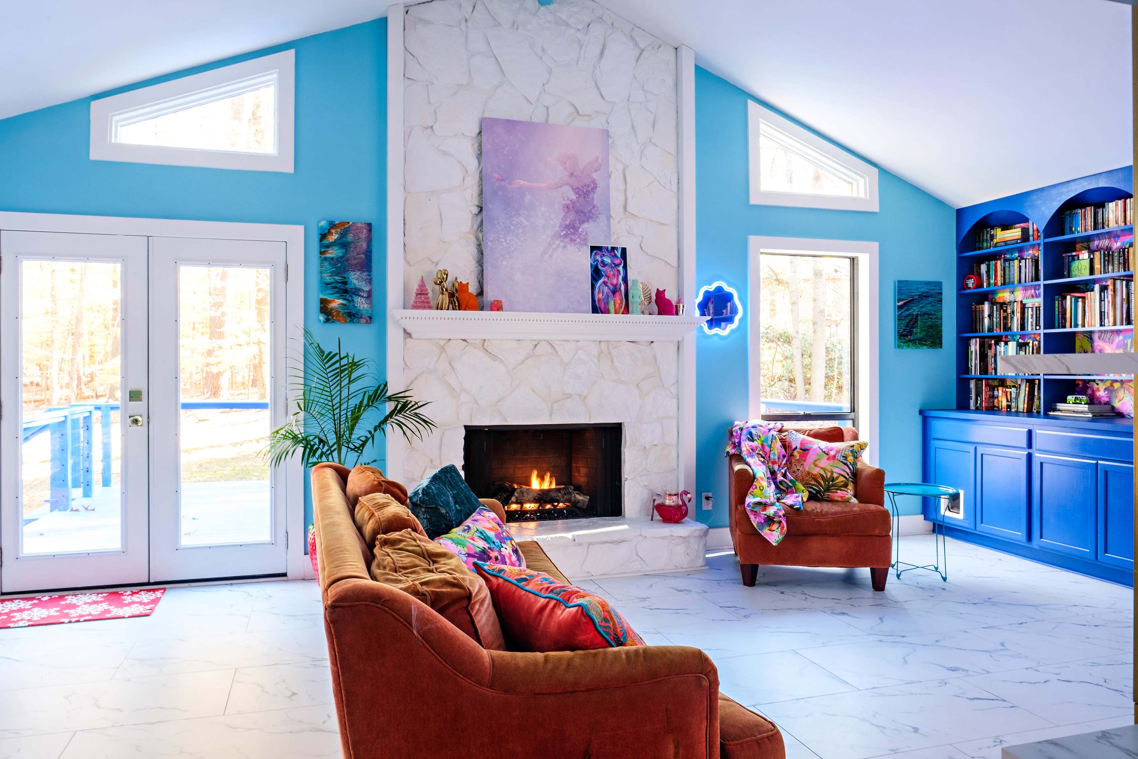 A cozy living room features a stone fireplace, a large window with a view, colorful decor, and a blue bookshelf filled with books.