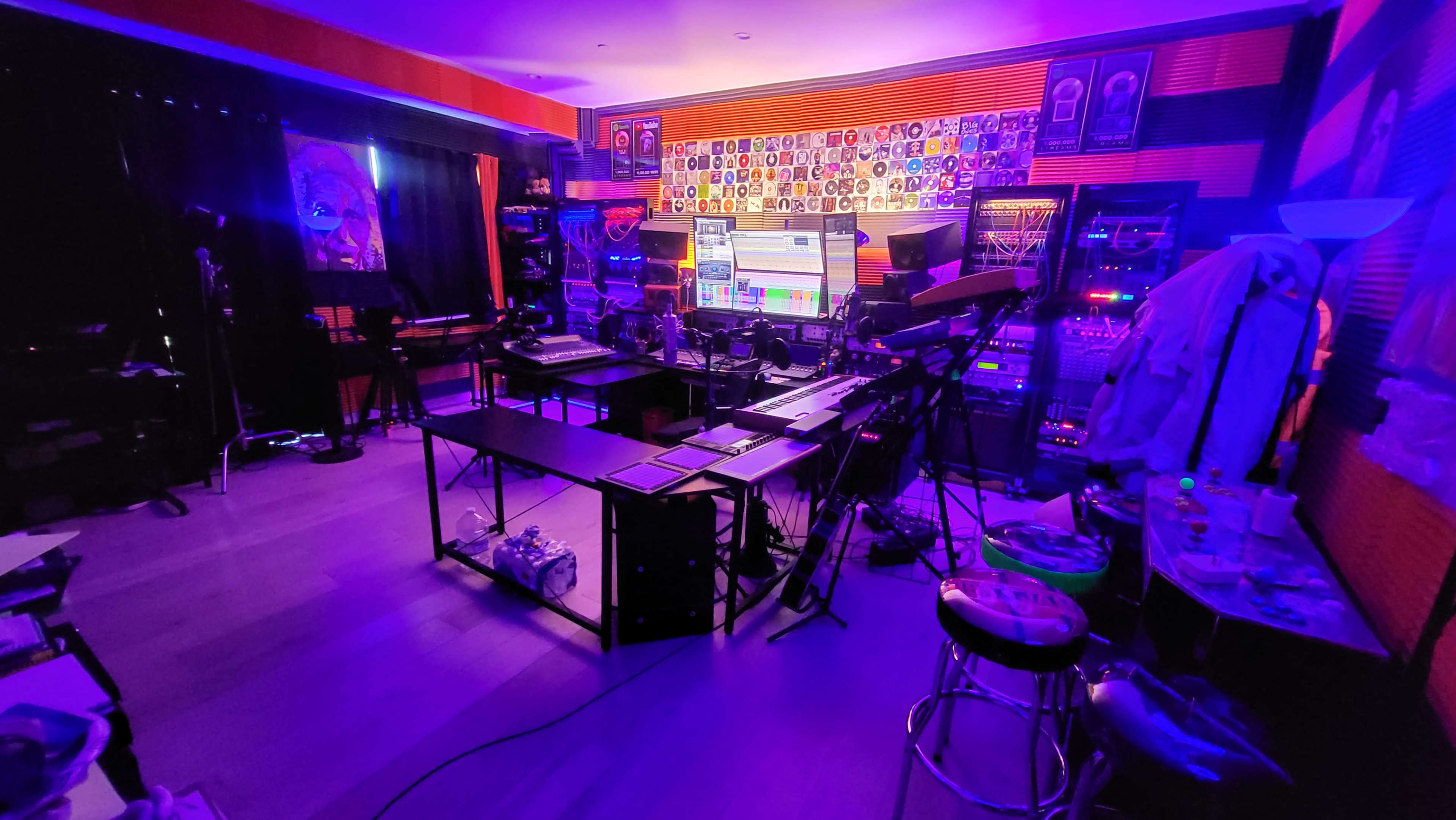 The image shows a dimly lit music studio filled with various instruments, equipment, and a large computer setup against a wall covered in colorful posters.
