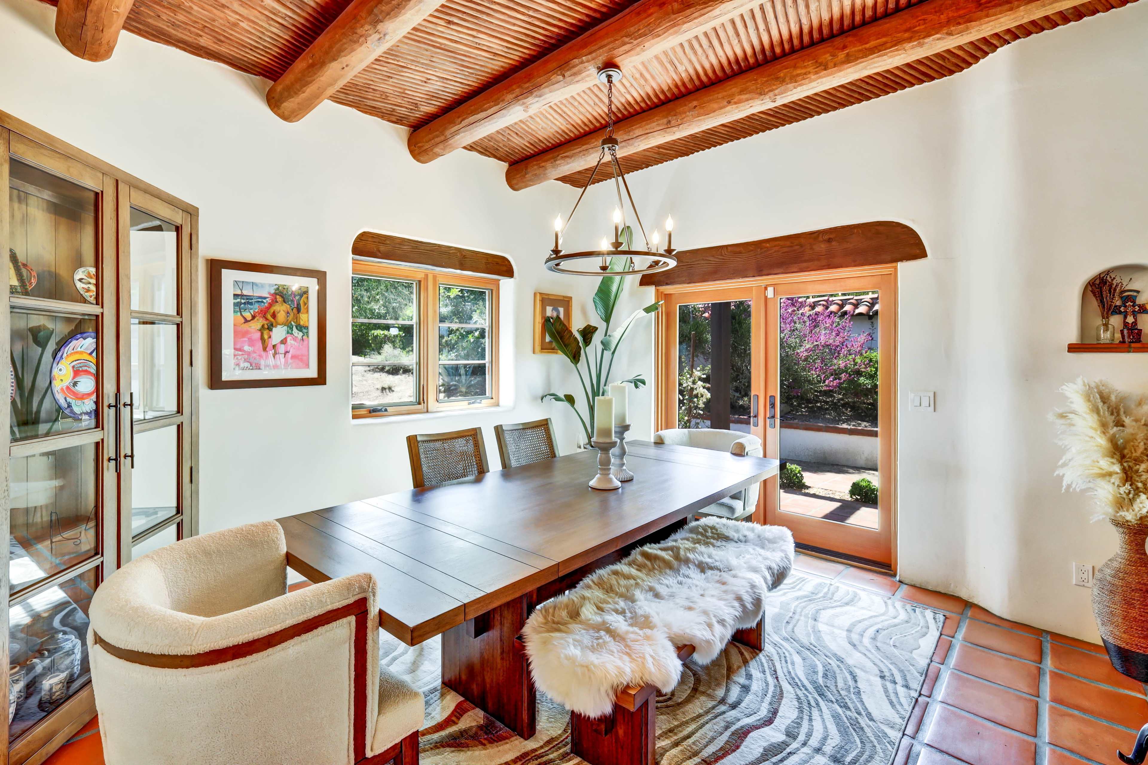 The image shows a dining room featuring a wooden table surrounded by upholstered chairs, with large windows and a sliding glass door leading to an outdoor space.