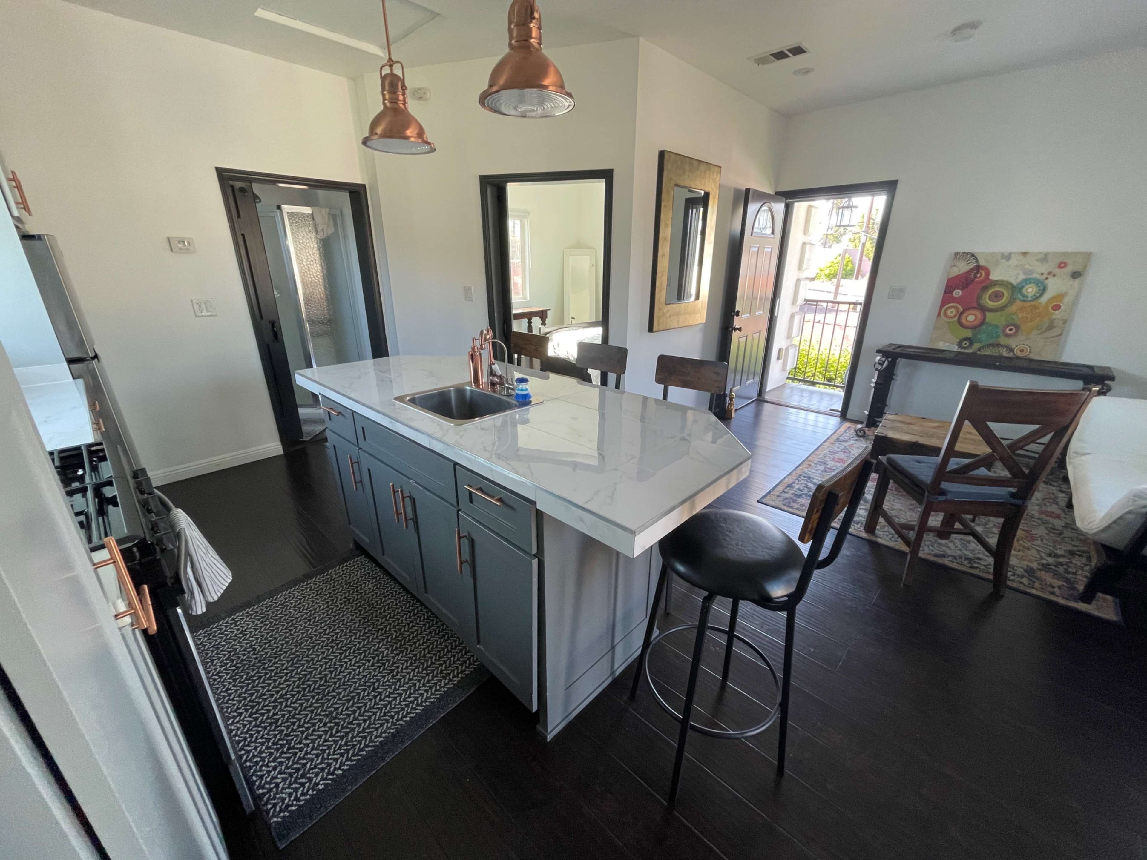 The image shows a modern kitchen with a marble countertop island, black bar stools, and a dining area with wooden chairs, featuring bright natural light.
