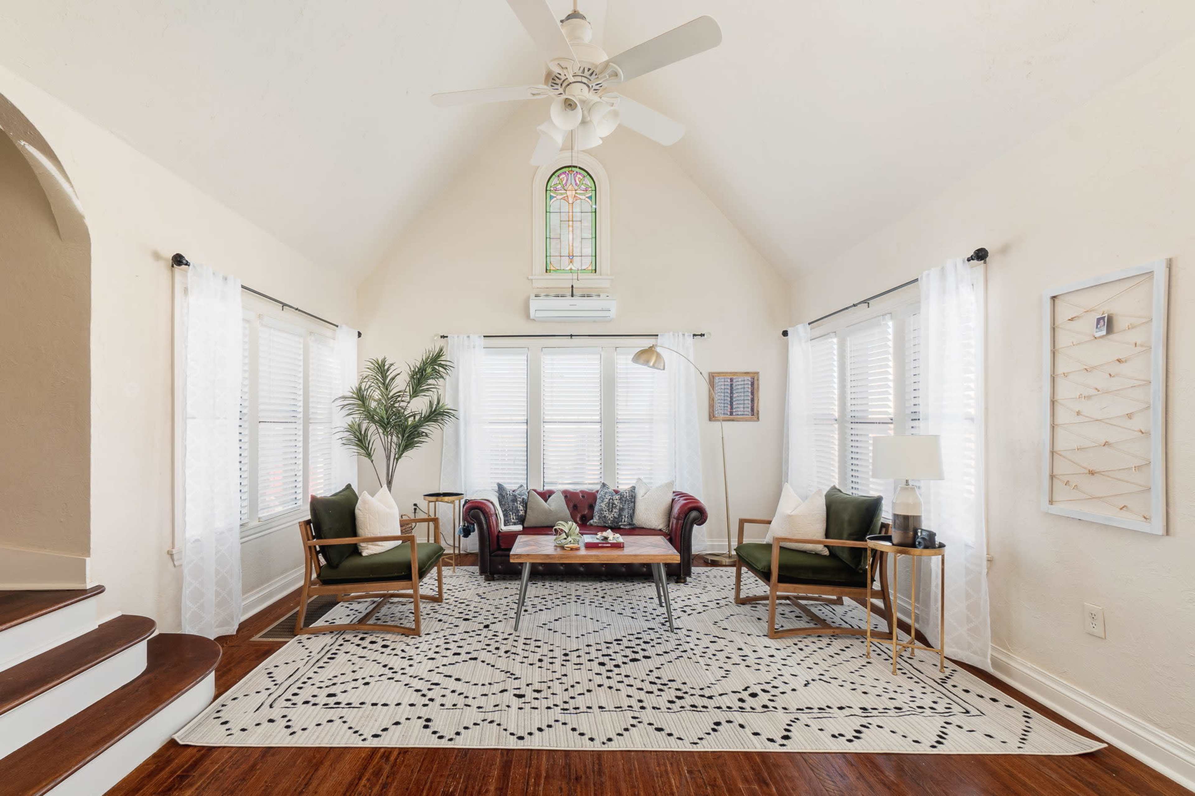 A bright living room with a high ceiling, featuring a red couch, green armchairs, a coffee table, and large windows adorned with light curtains.