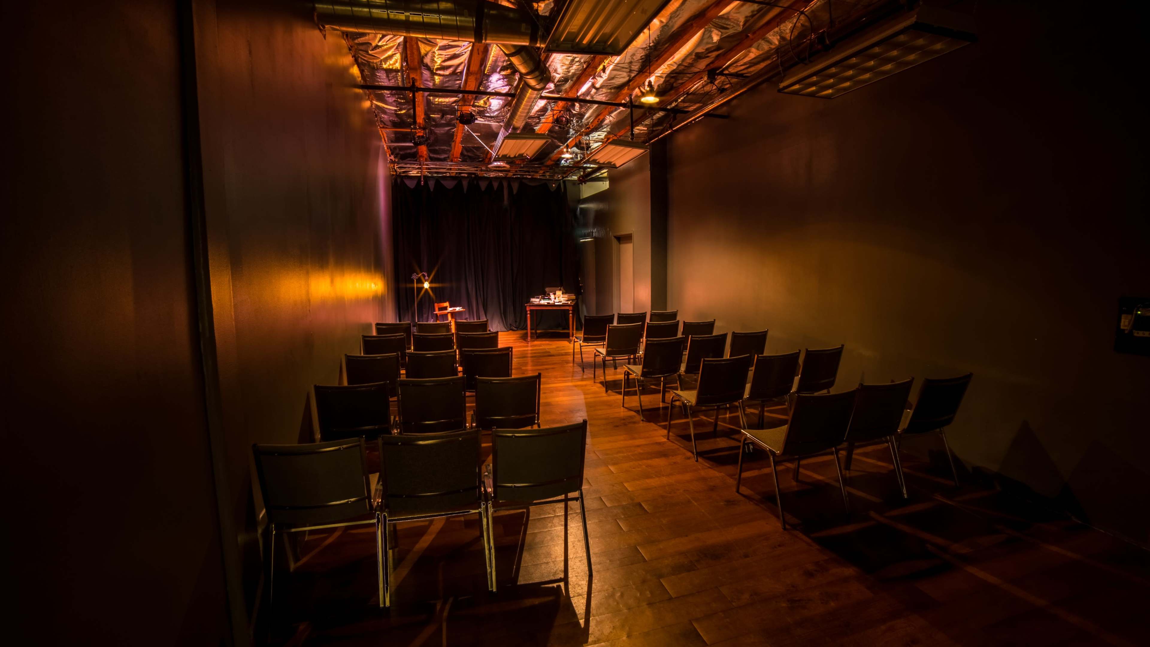 The image shows a dimly lit auditorium with rows of metal chairs facing a small stage and a table at the front.