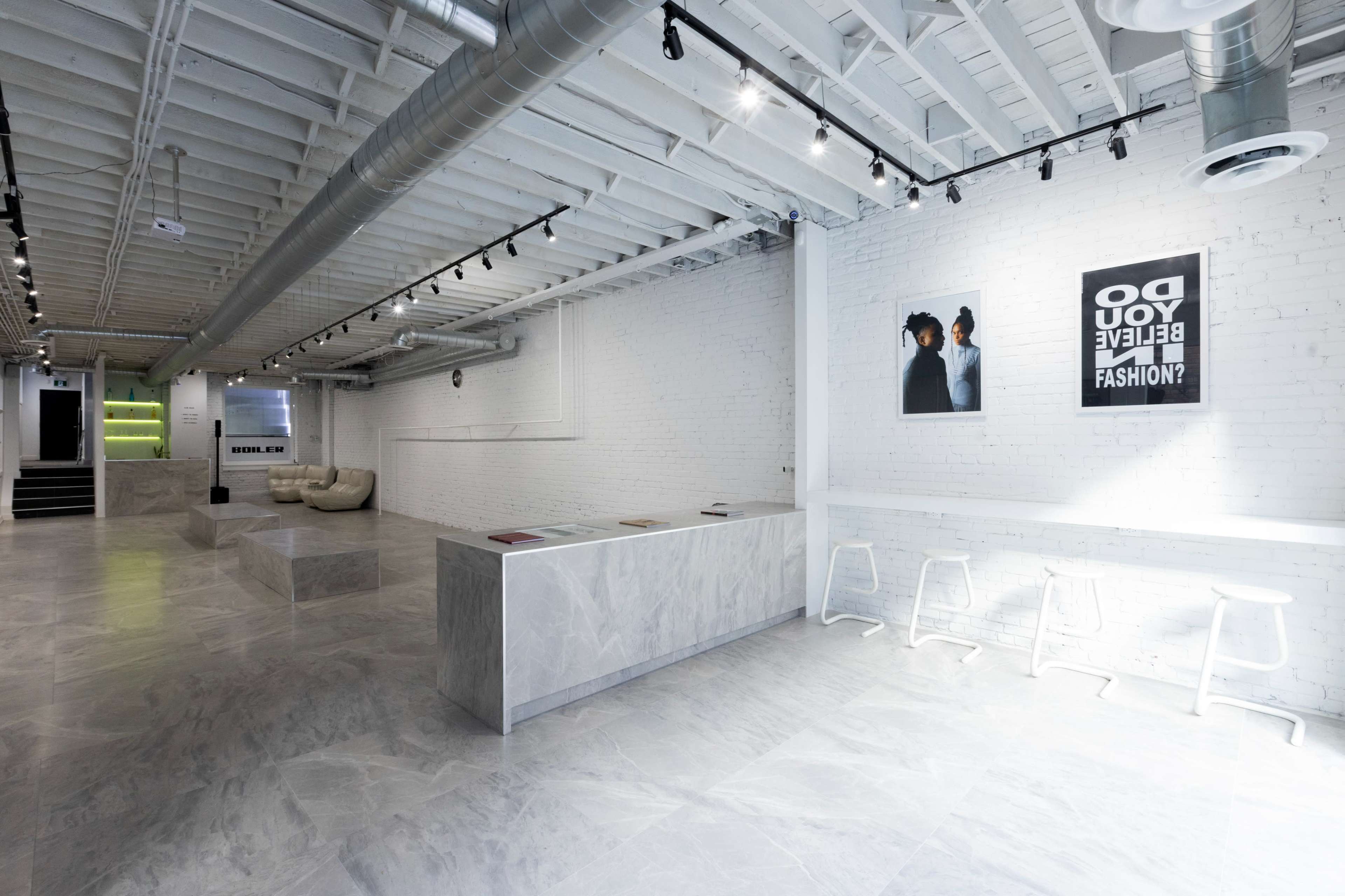 The image shows a modern, minimalist interior of a fashion showroom with white brick walls, a marble reception counter, and seating areas, illuminated by overhead lights.