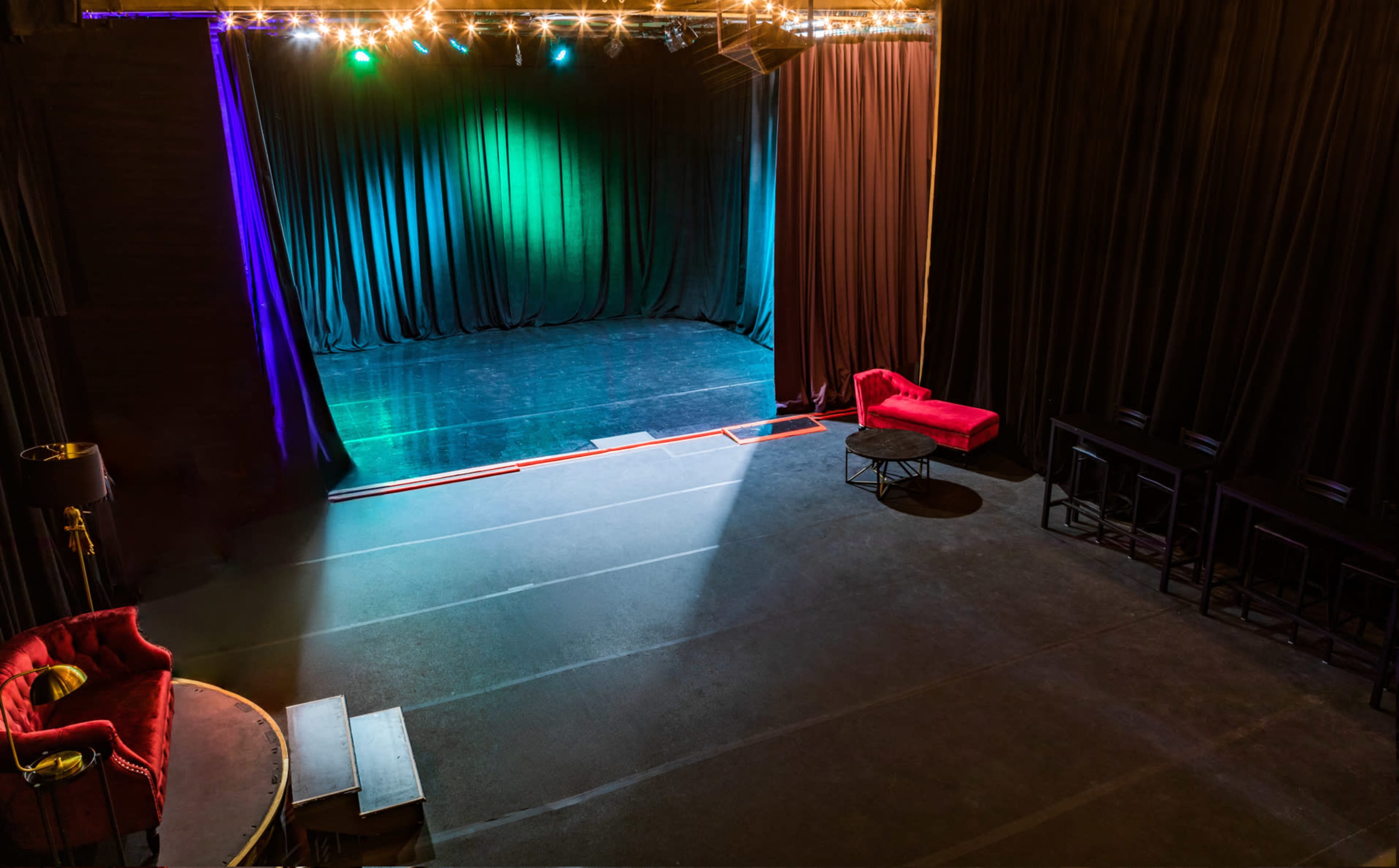 The image shows an empty theater stage with green and blue lighting, surrounded by black curtains, and a seating area with red couches and a circular table.