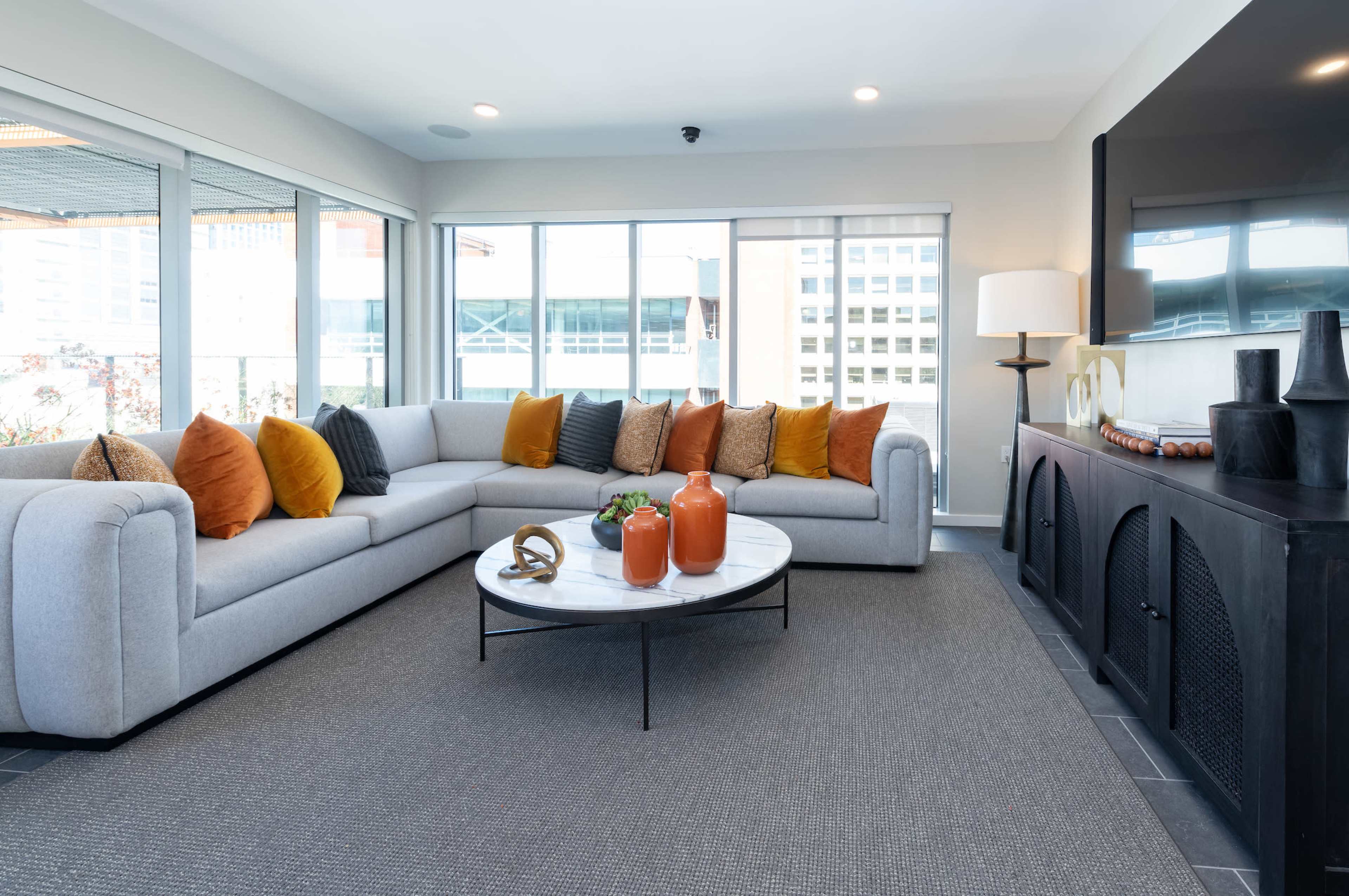 A modern living room with a large gray sectional sofa adorned with colorful pillows, a circular coffee table, and large windows providing natural light.