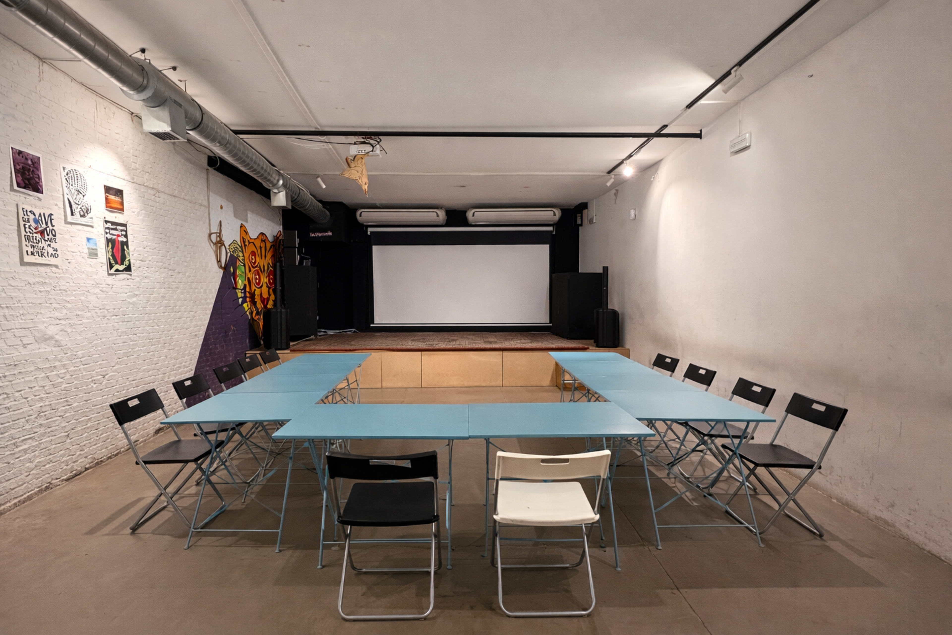 A rectangular arrangement of tables and chairs faces a blank screen in a room with white walls and a wooden stage.