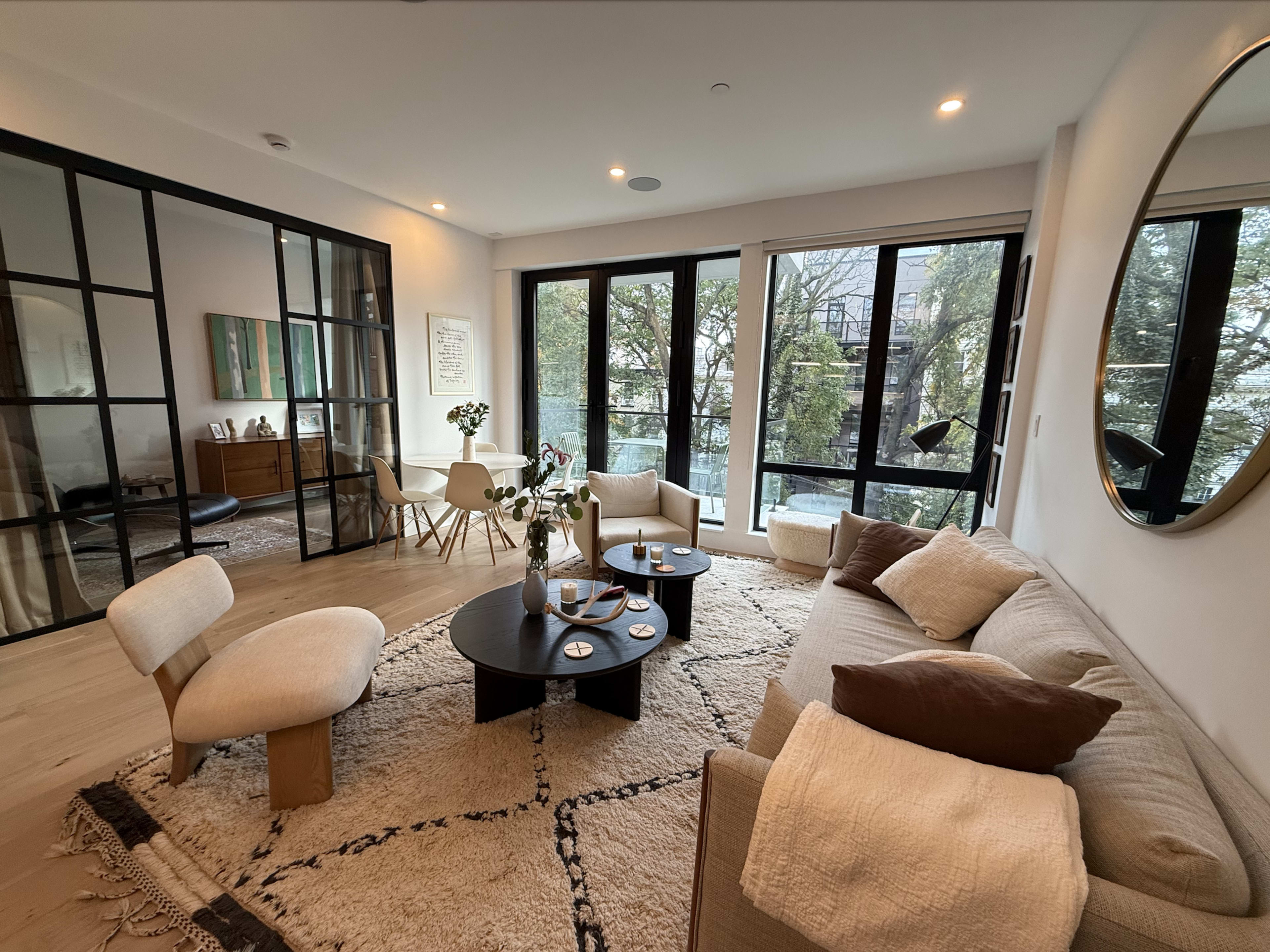 The image shows a modern living room with a light-colored sofa, a round coffee table, and large windows that offer a view of greenery outside.