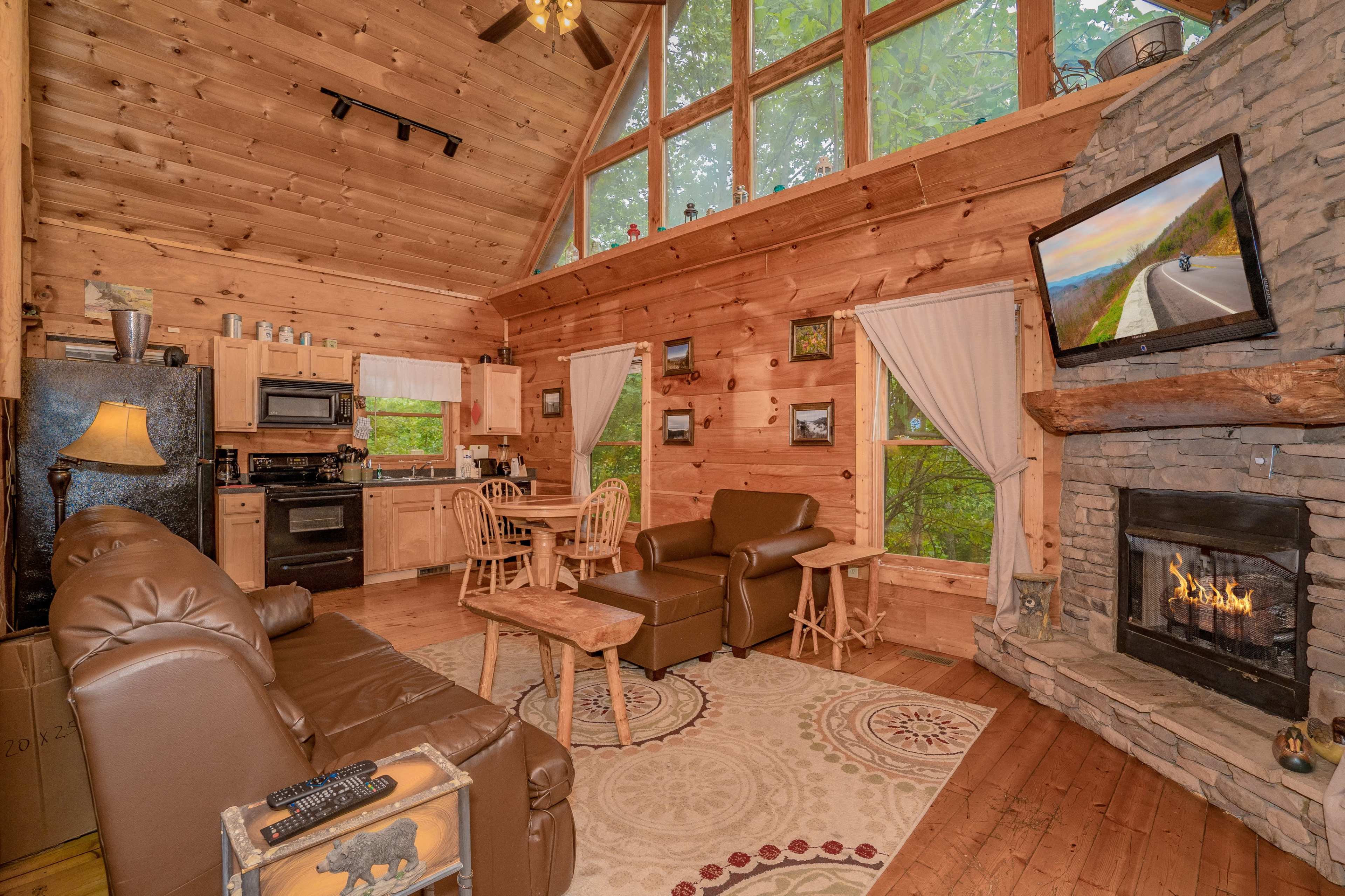 The image shows a cozy cabin interior with wooden walls, a stone fireplace, a kitchen area, and large windows allowing natural light.