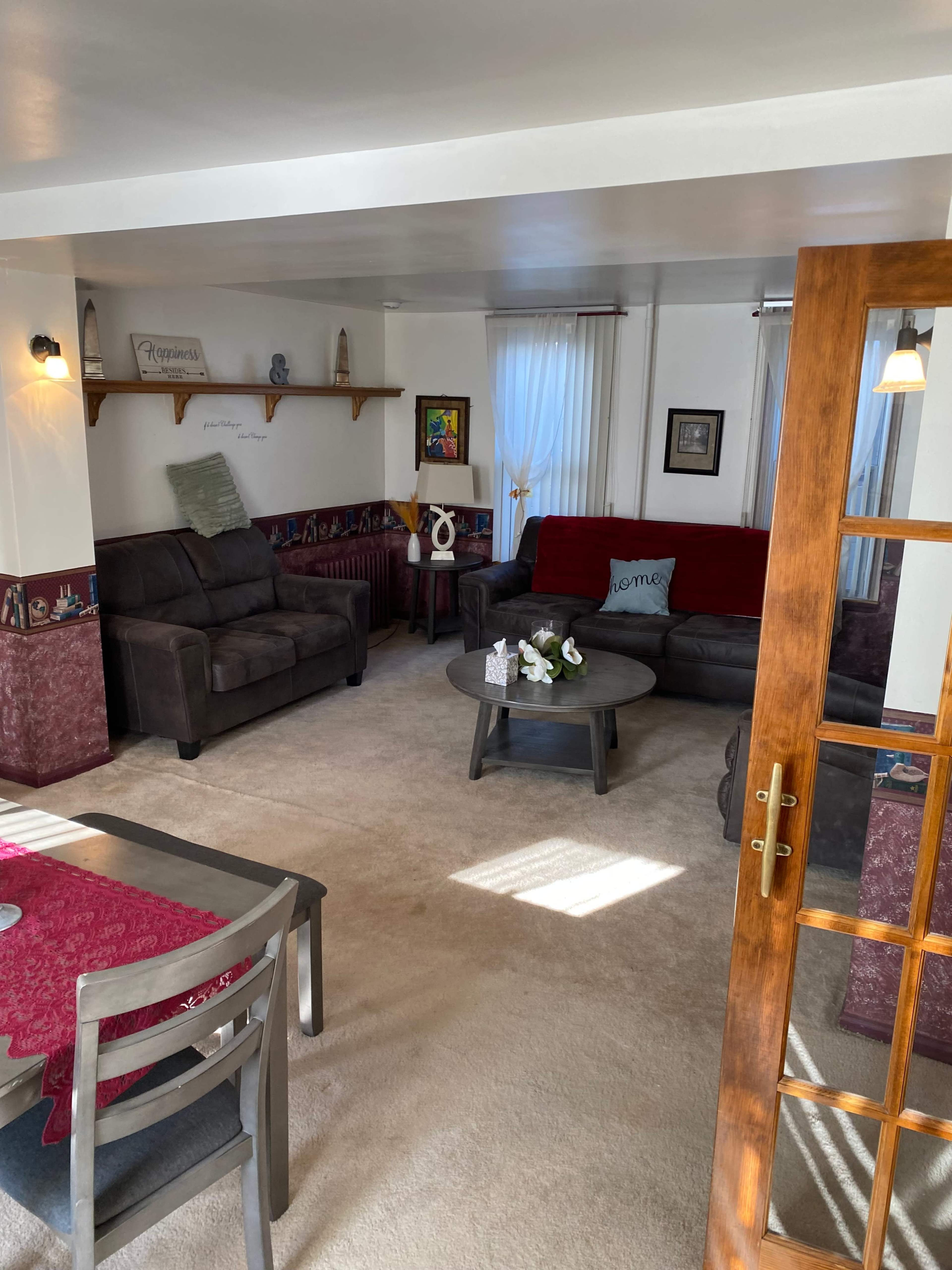 The image shows a living room with a red couch, two gray sofas, a coffee table, and a dining area with a table set for meals.