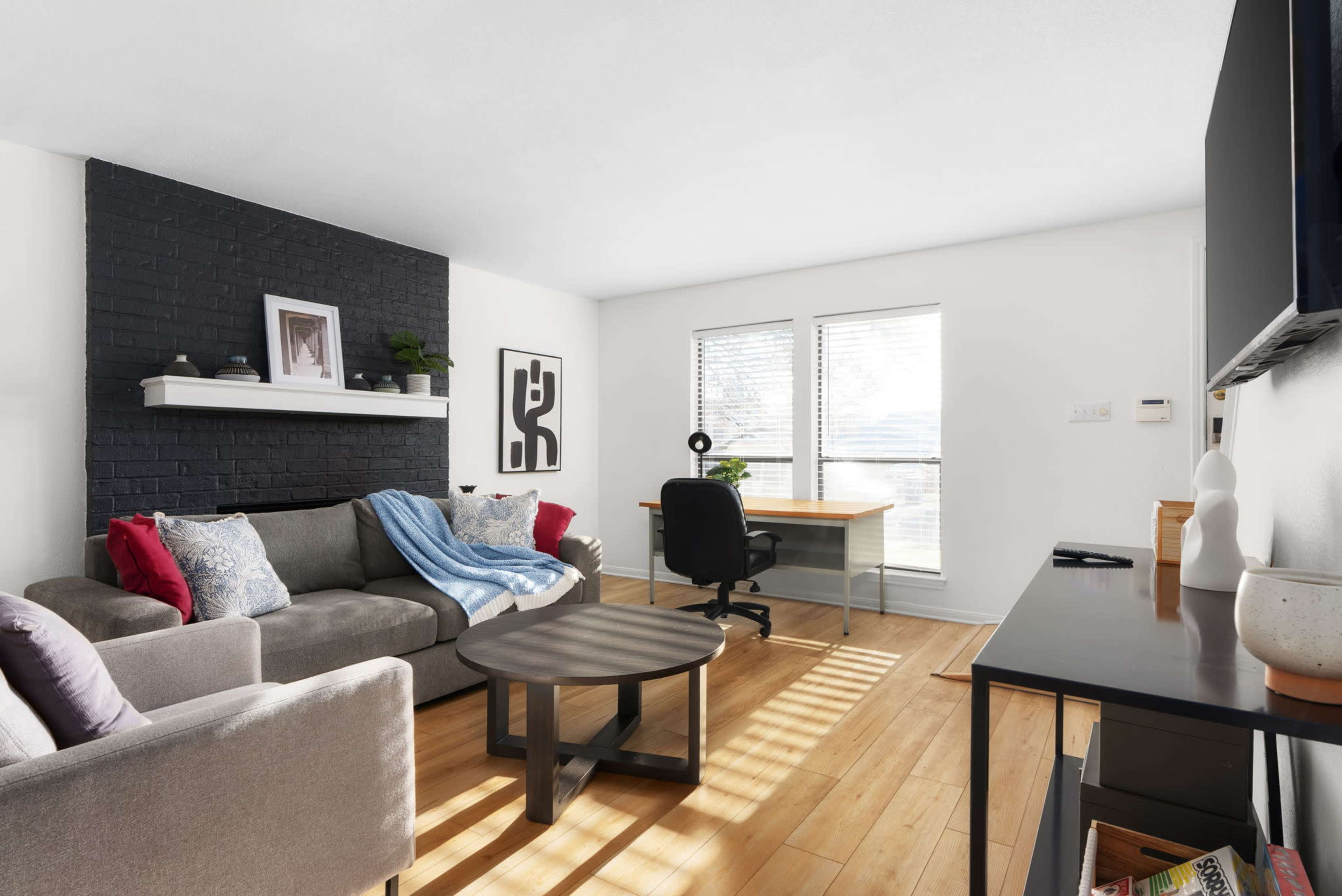 A living room features a gray sofa with decorative pillows, a round coffee table, a desk with a chair, and large windows allowing natural light.