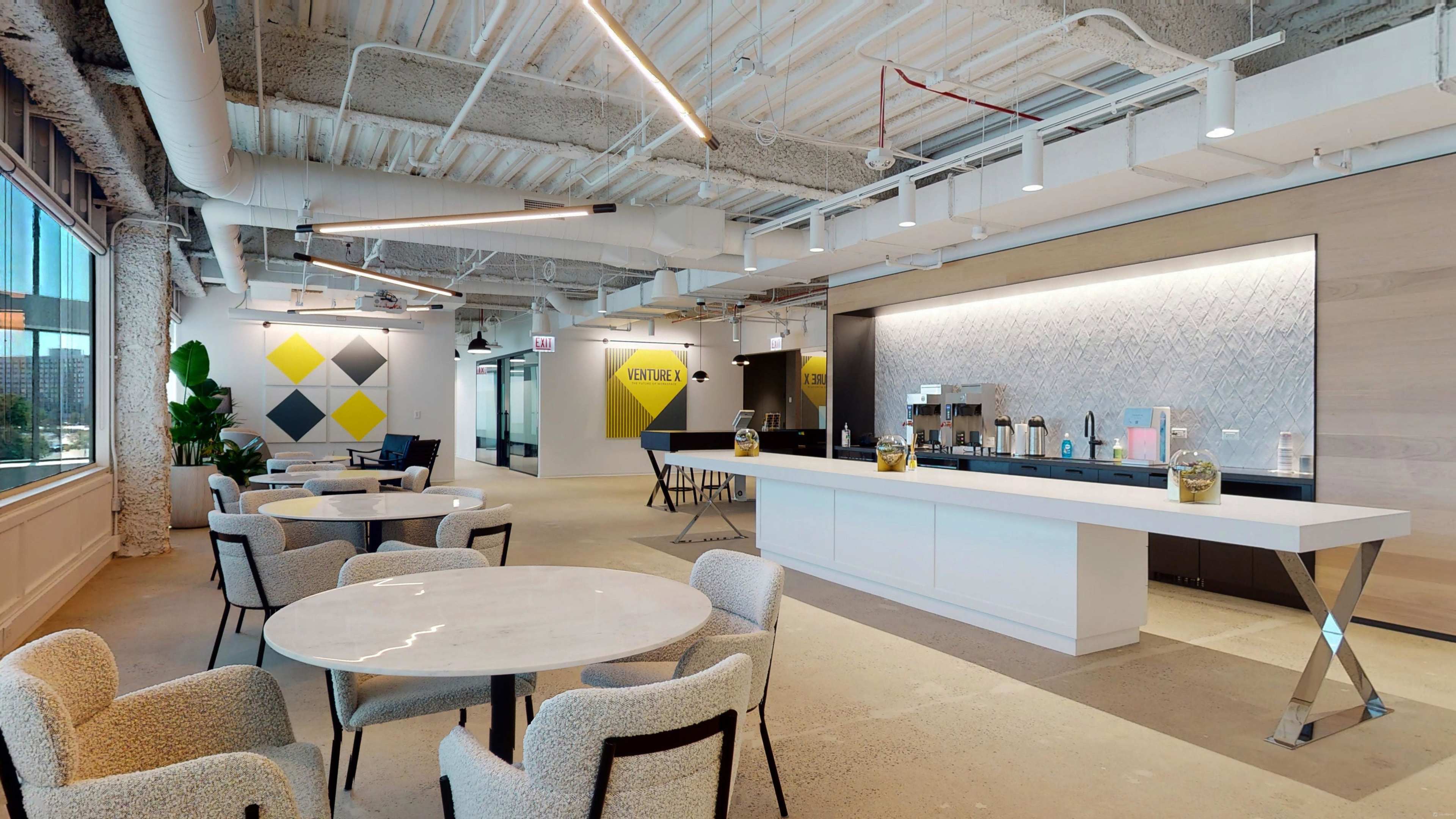 A modern office space features a bright area with a coffee bar, tables, and geometric wall decorations.