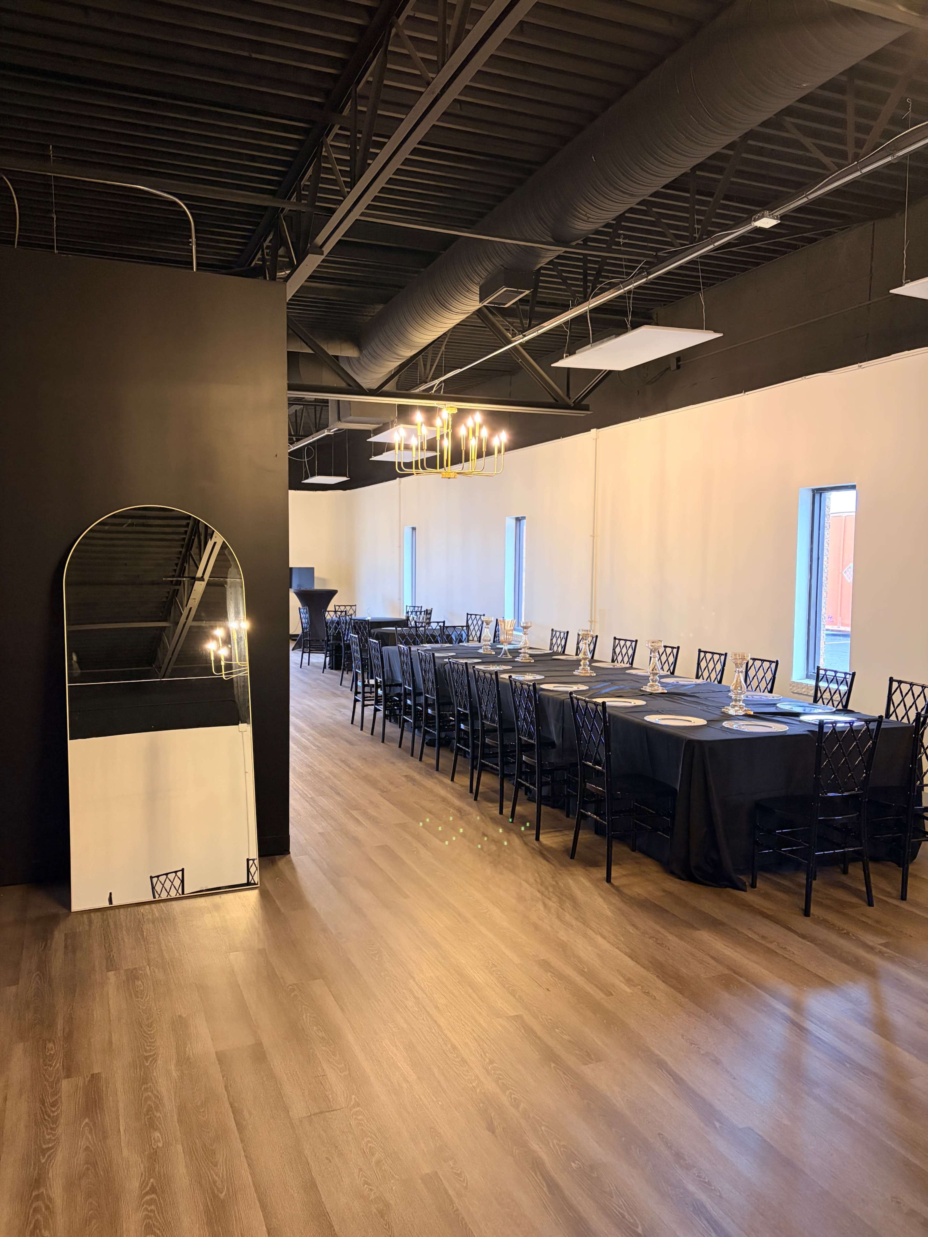 A spacious dining area features a long table with black tablecloths and elegant chairs, illuminated by modern chandeliers, alongside a wall with a large mirror.