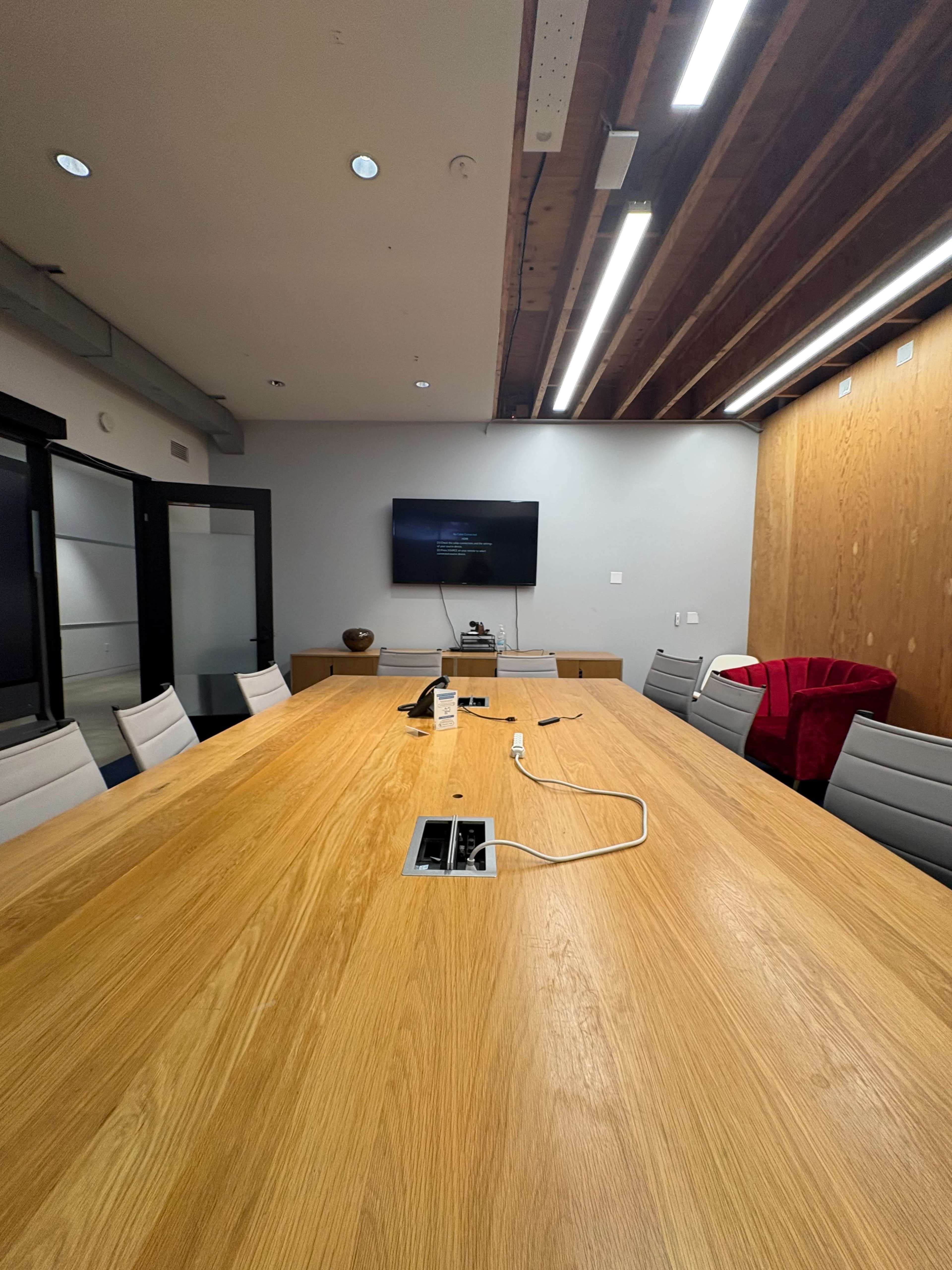 A large conference room features a wooden table surrounded by gray chairs, with a wall-mounted television and a wooden cabinet in the background.