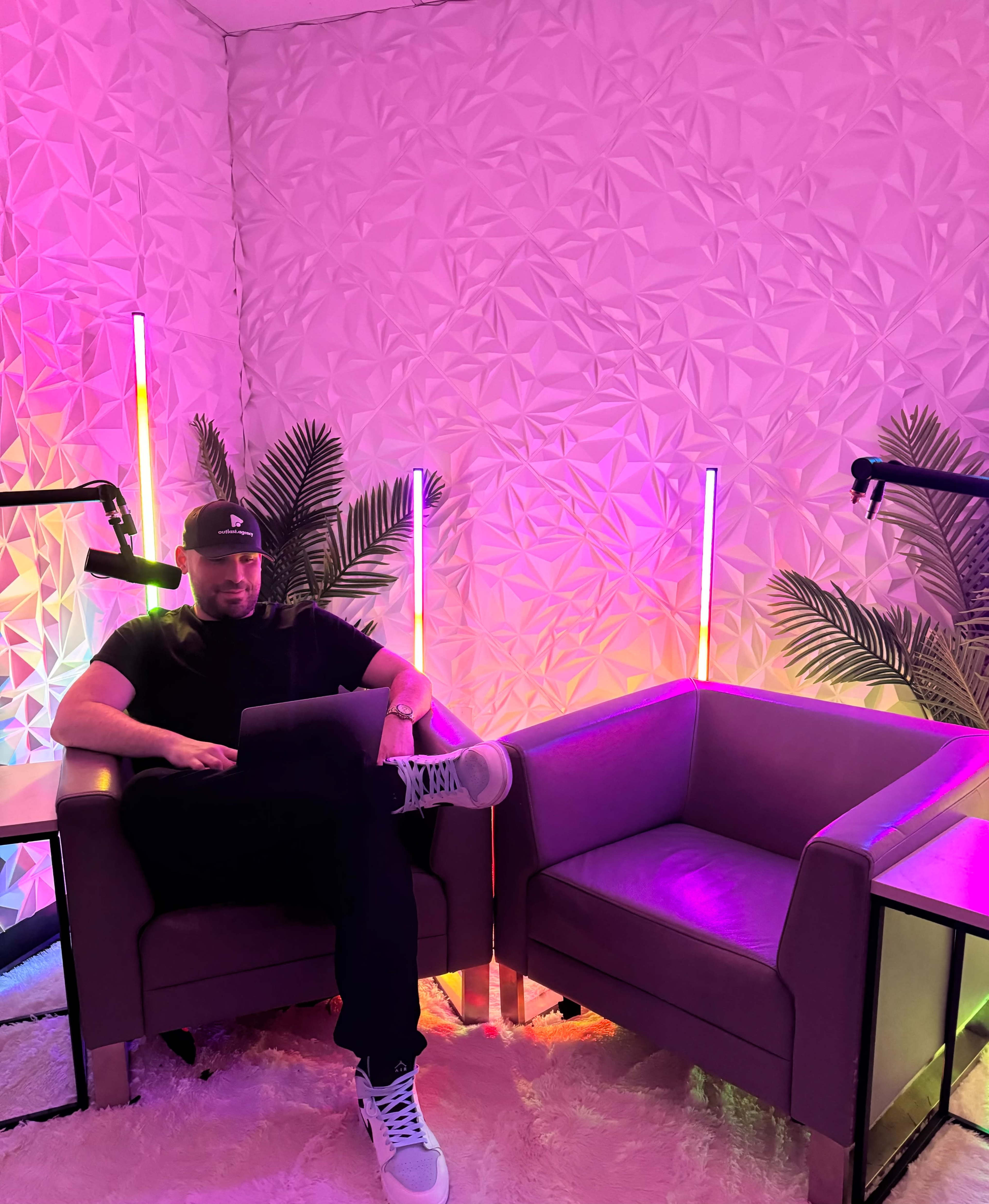 A man sits on a chair in a brightly lit room with geometric patterned walls and plants, using a laptop.