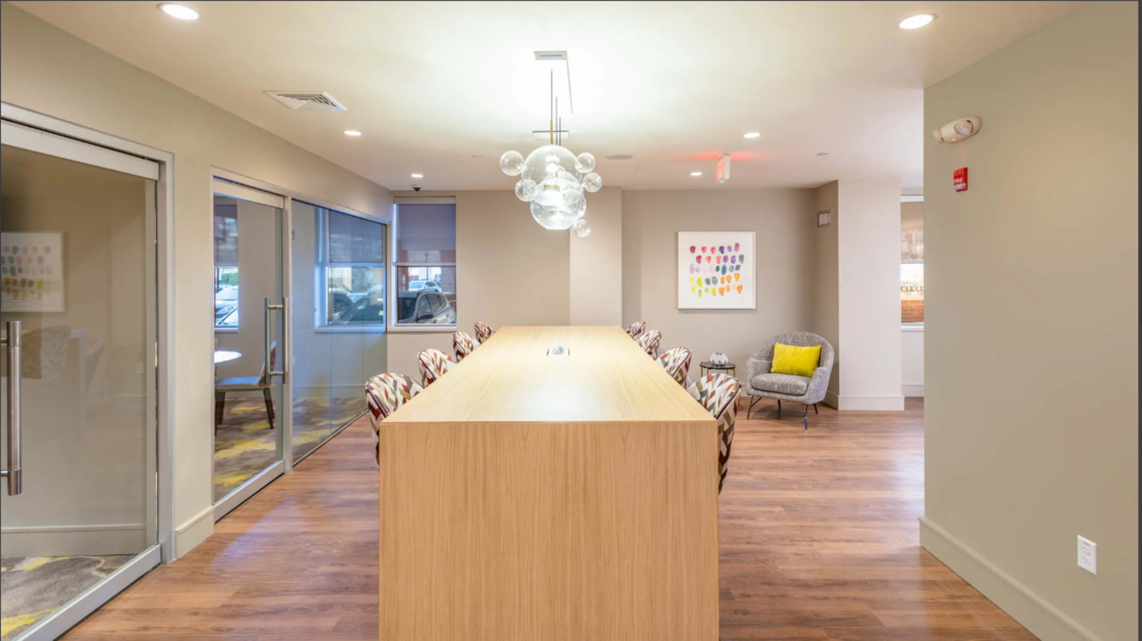 A modern office space features a long wooden table with seating and a seating area with a gray chair and colorful artwork on the wall.