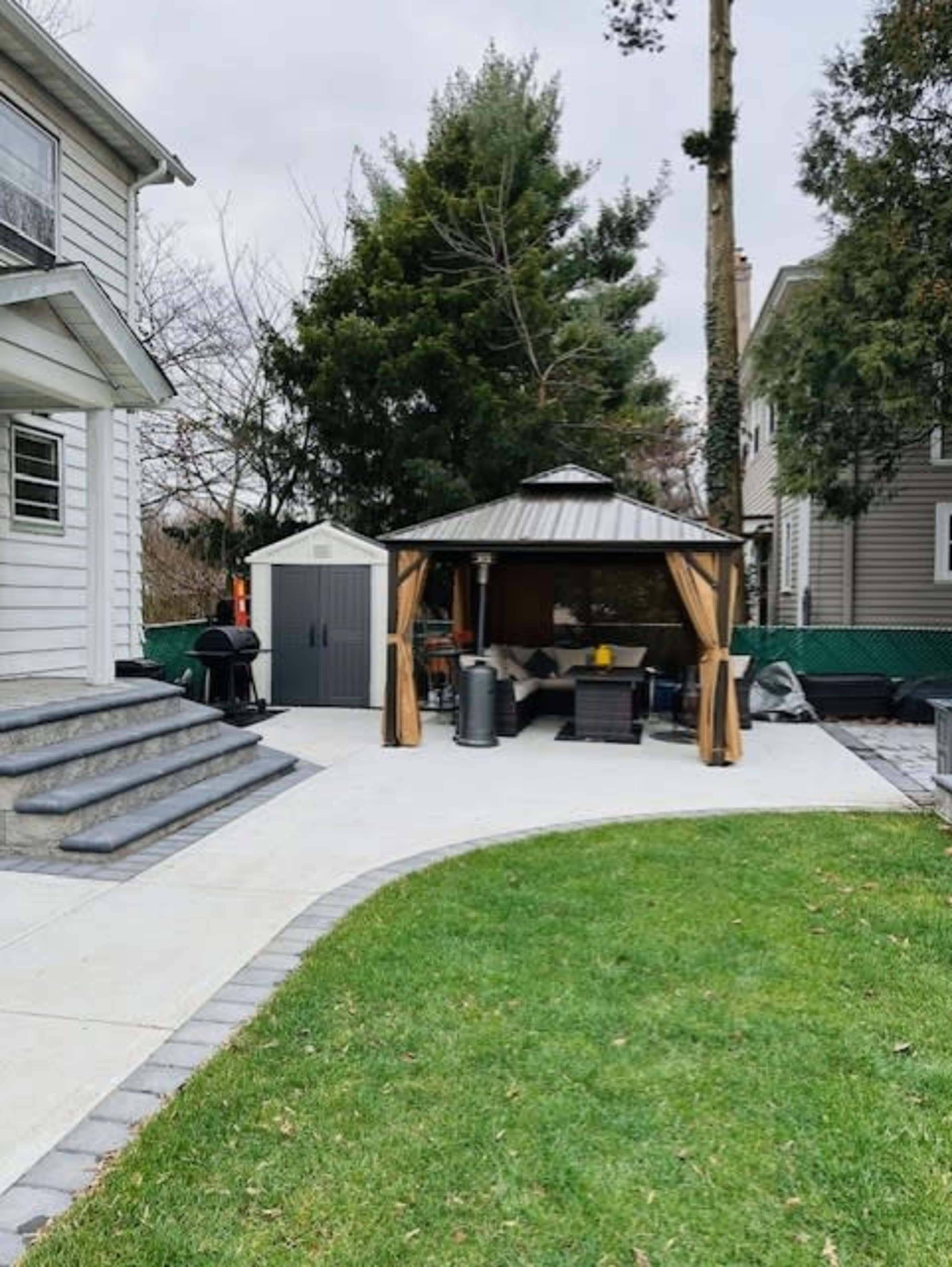 A backyard patio featuring a gazebo with curtains, a grill, and a garden shed, all surrounded by grass and trees.