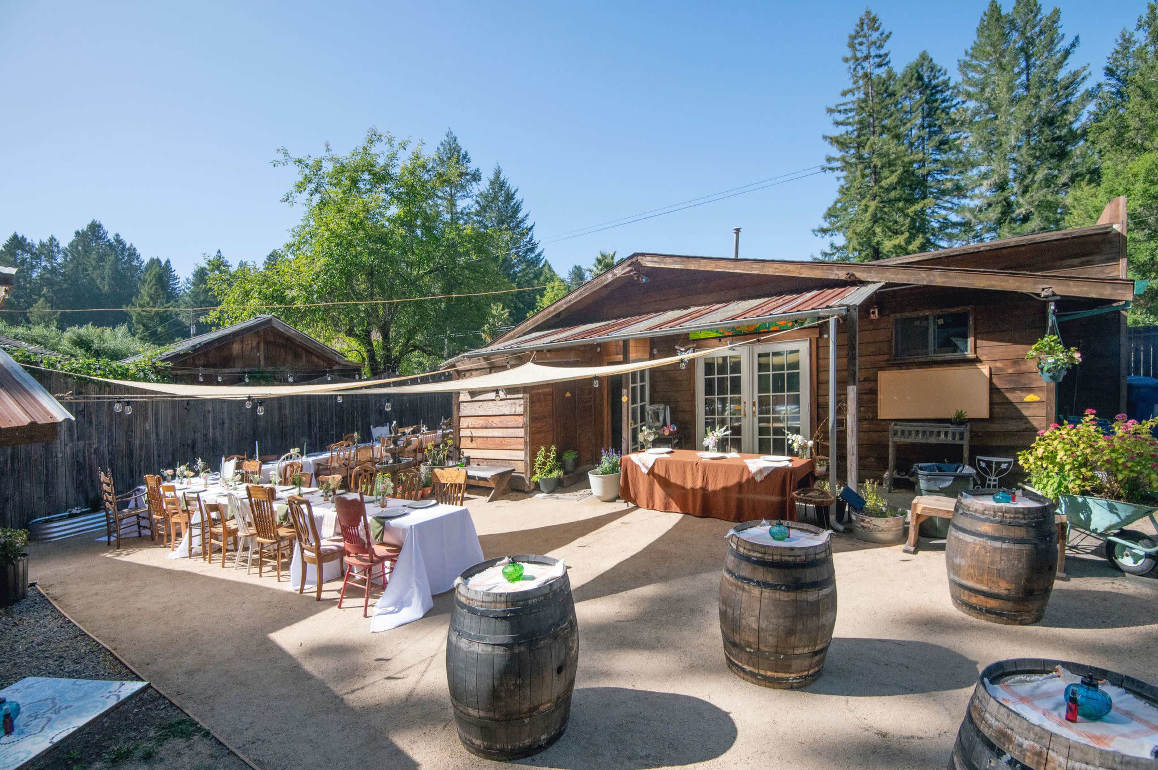The image shows a rustic outdoor event space with wooden structures, tables set with white linens, and barrels arranged throughout the area.