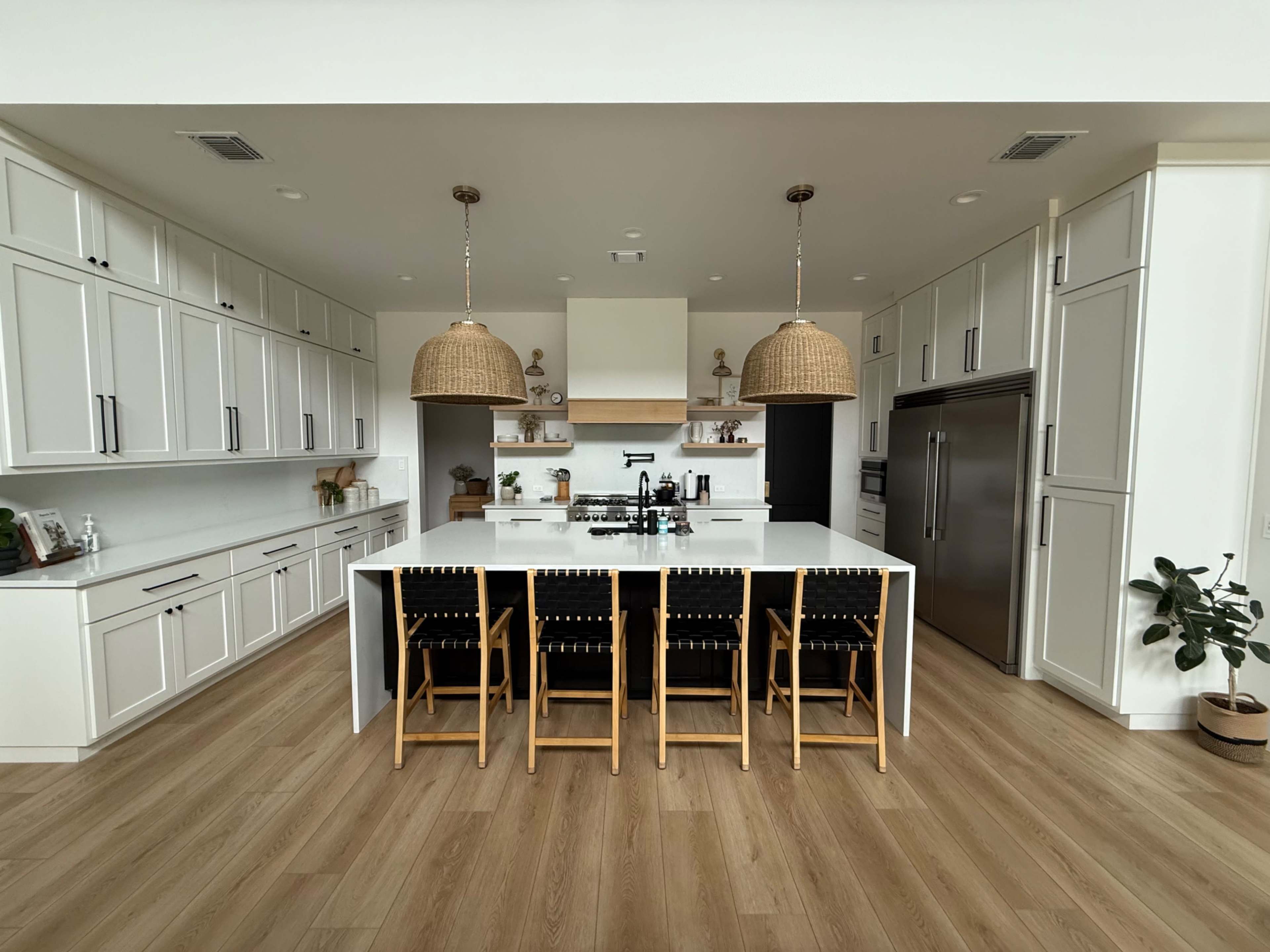 The image shows a modern kitchen with white cabinetry, a large island featuring a sink, and four wooden bar stools.