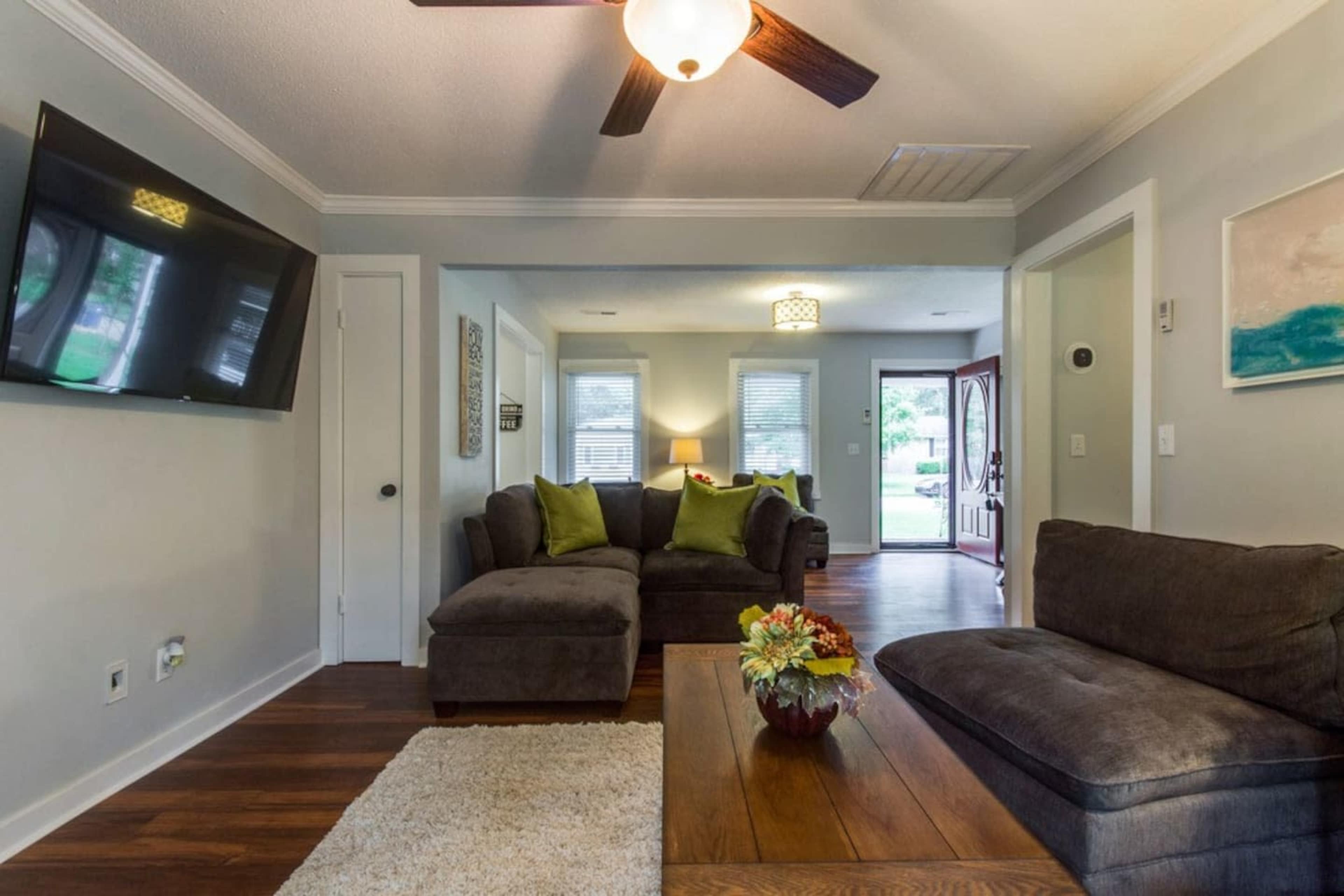 The living room features a sectional sofa and a coffee table, with a television mounted on the wall and a front door visible in the background.