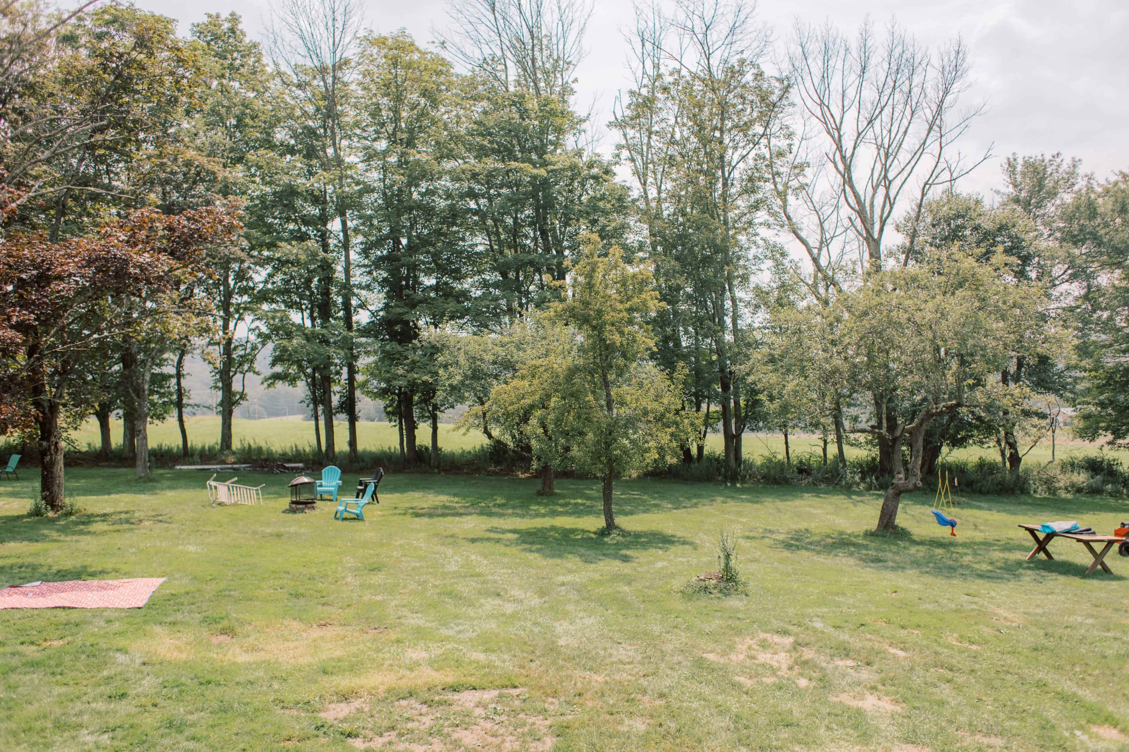 The scene depicts a grassy yard with a few trees, outdoor seating, a swing set, and a picnic area under a clear sky.