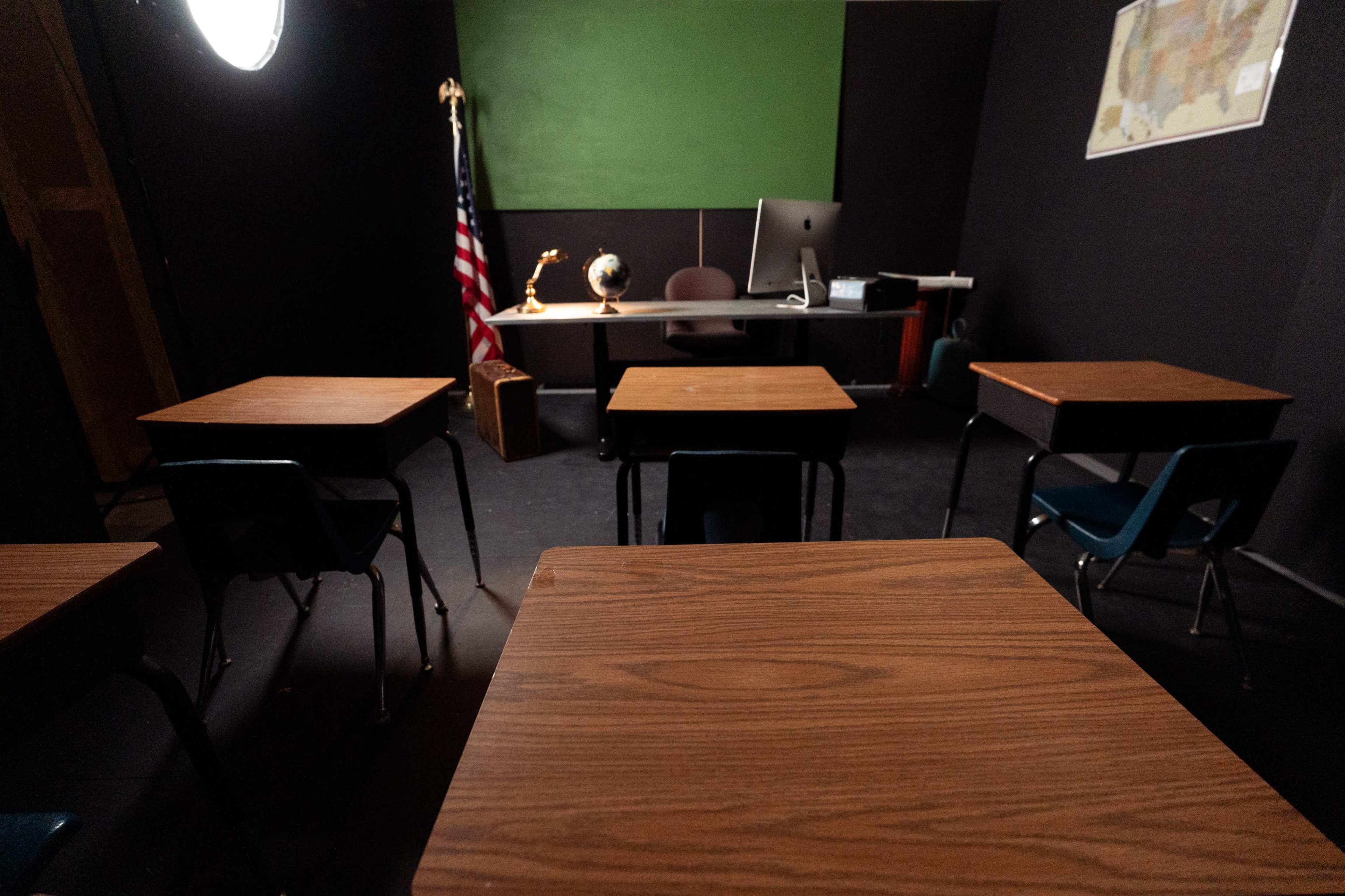The image shows a classroom setting with several wooden desks facing a teacher's desk, which is accompanied by an American flag, a globe, and a map on the wall.