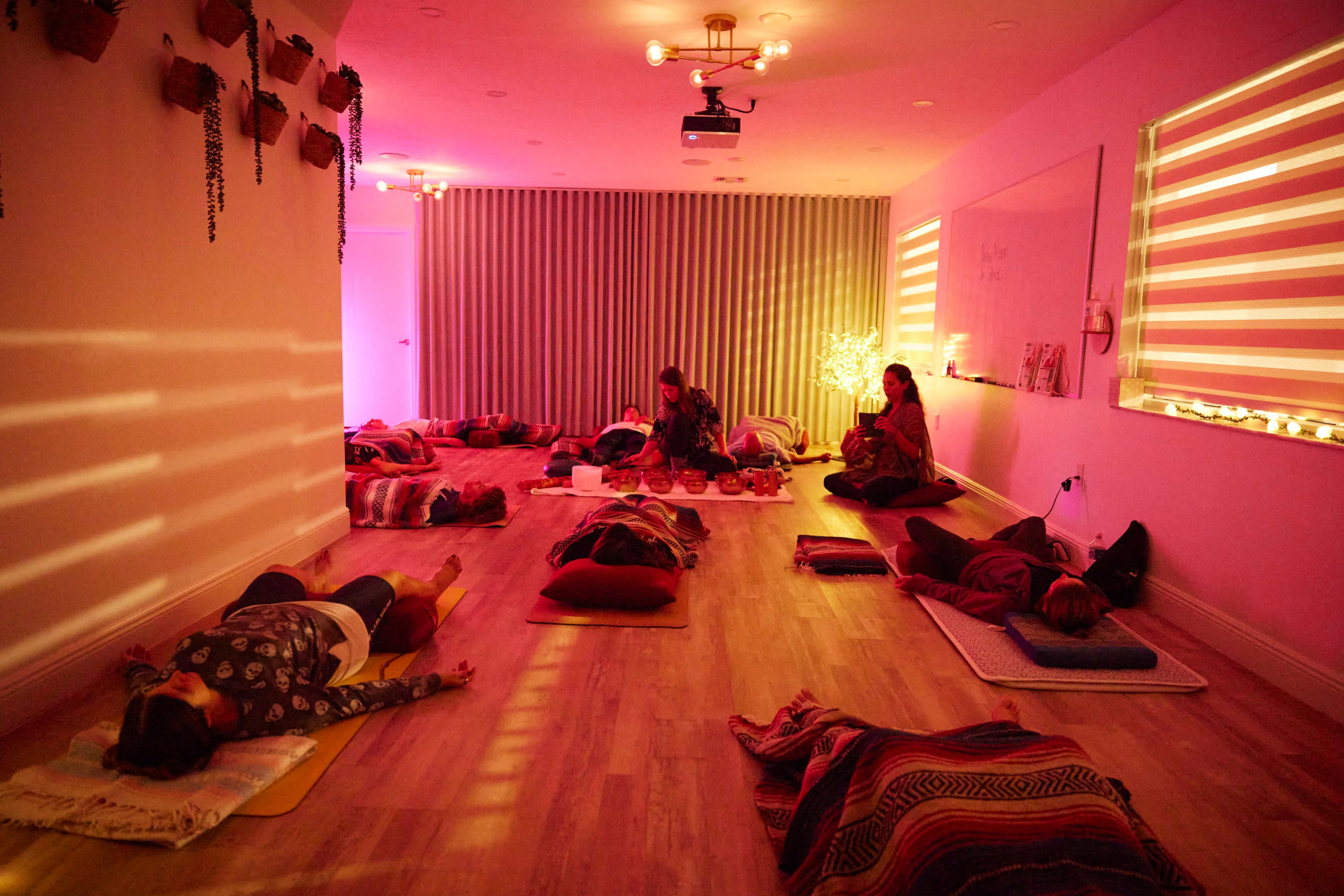 The image shows a tranquil yoga studio filled with people practicing relaxation on mats, with soft lighting and decorative walls.