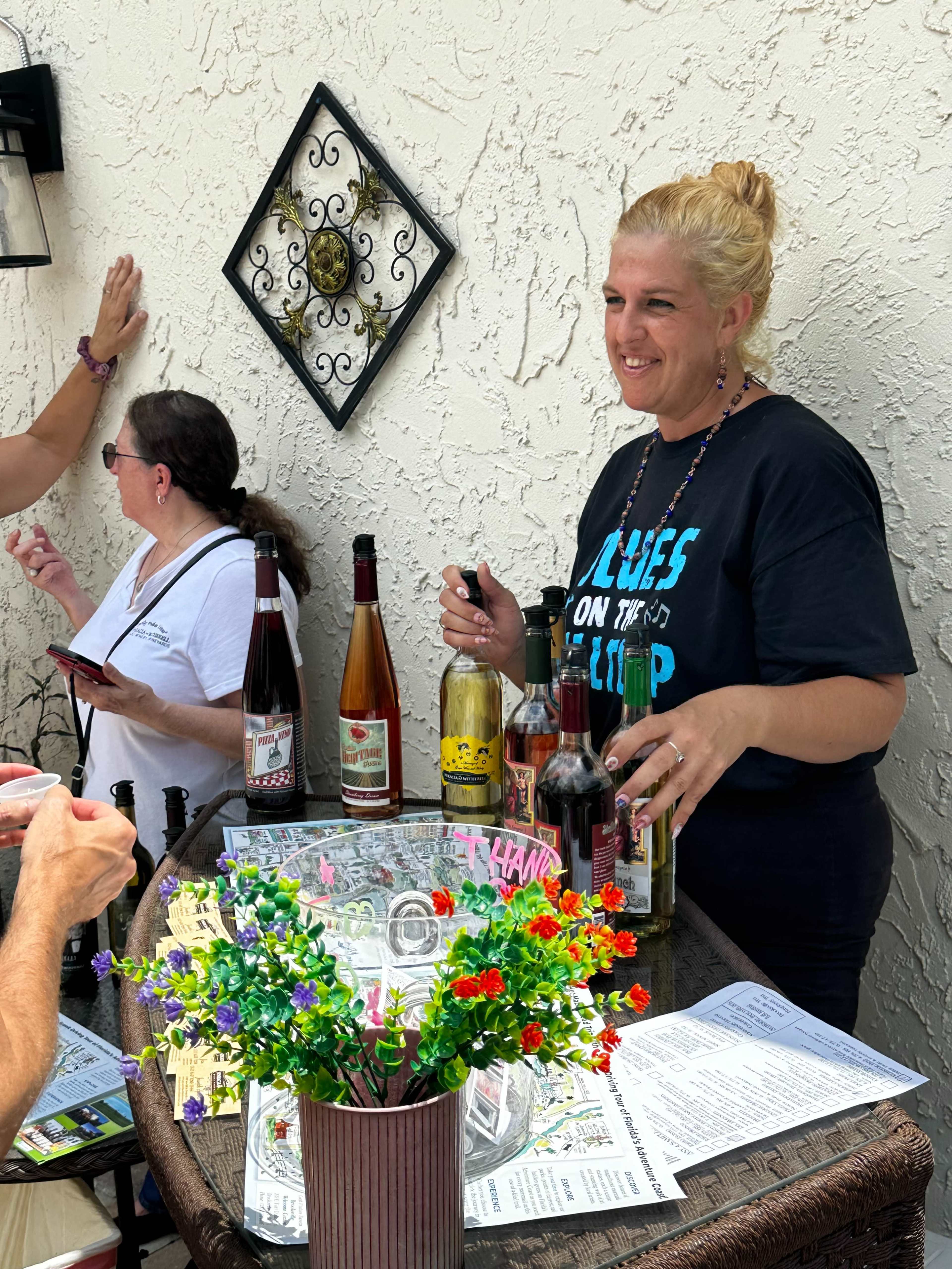 A woman stands behind a bar cart with various bottles of wine and spirits, while another person looks on in the background.